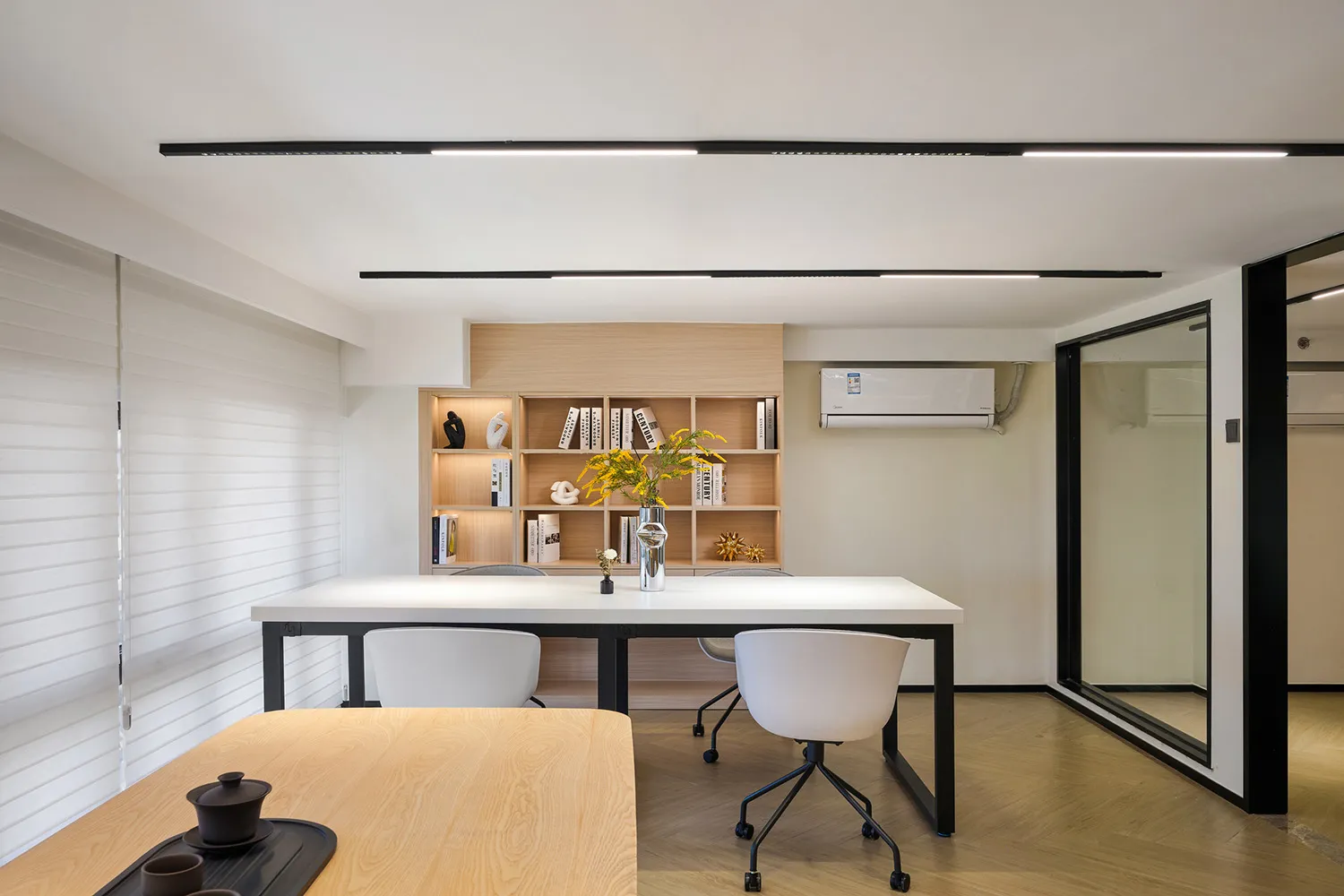 Modern minimalist home office with white rectangular table, white ergonomic chairs, light oak built-in bookshelf, and black linear ceiling lights.
