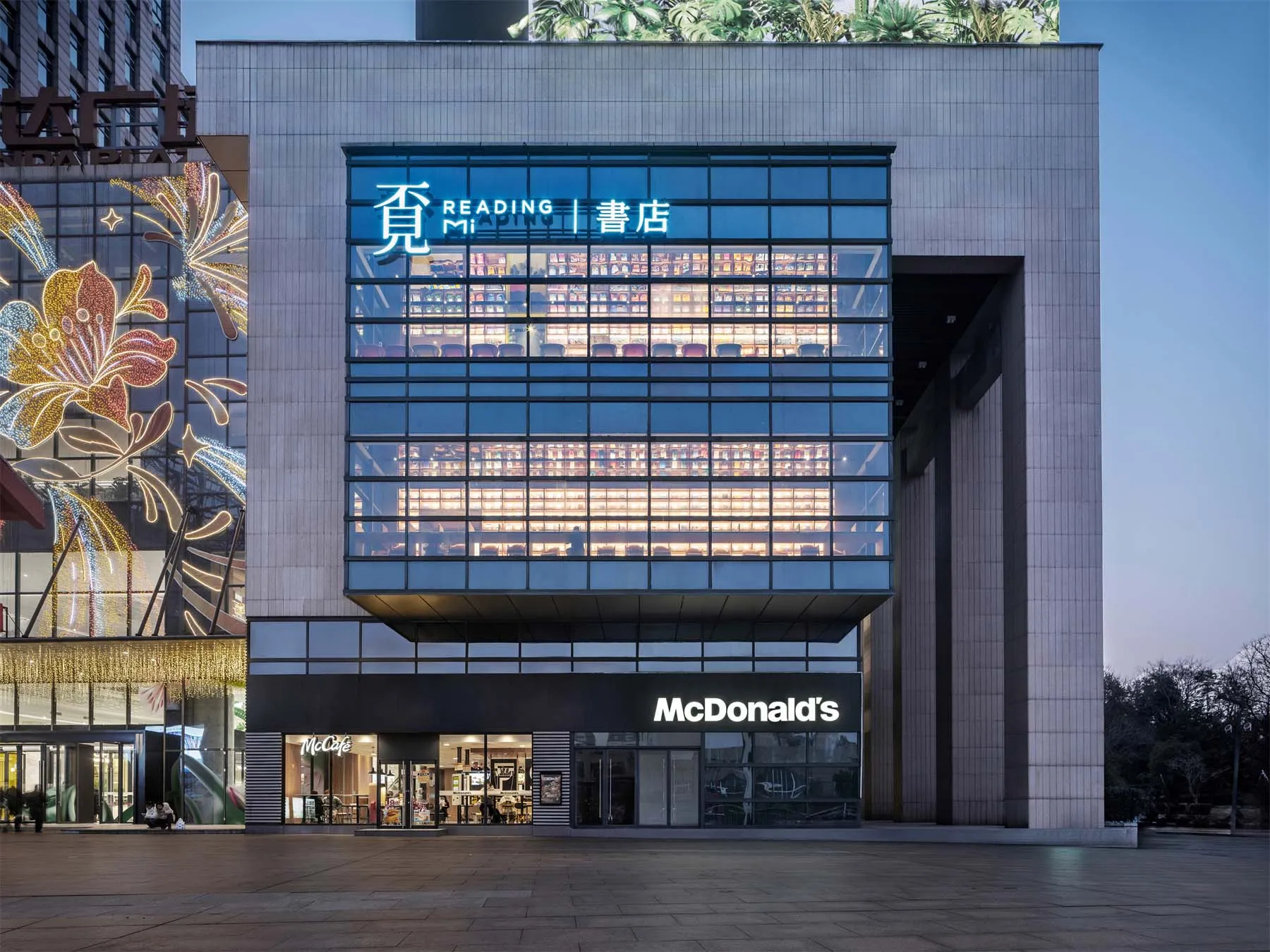 Night view of a modern commercial building featuring a glowing double-height glass reading room with bookshelves and the Reading Mi sign, positioned above a McDonald’s restaurant with festive floral light installations on the adjacent structure.