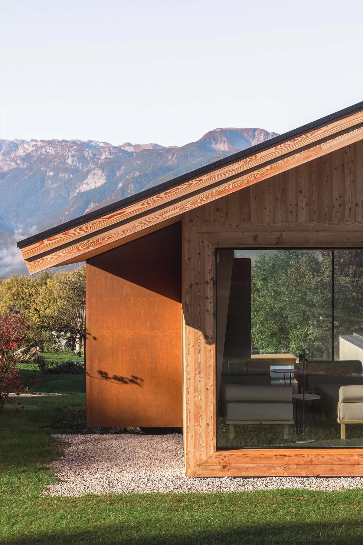Exterior view of a modern wooden cabin featuring a large Corten steel panel, expansive glass windows revealing the living area, set against a scenic mountain range with morning mist.