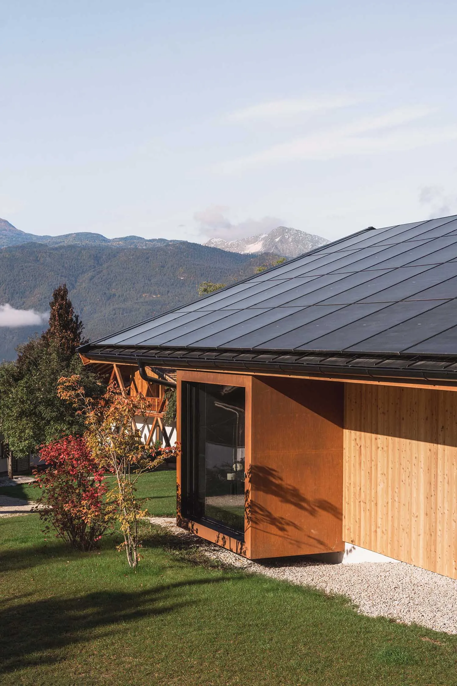 Modern mountain cabin featuring corten steel panels and light wood cladding with large windows overlooking snow-capped peaks and autumn foliage.