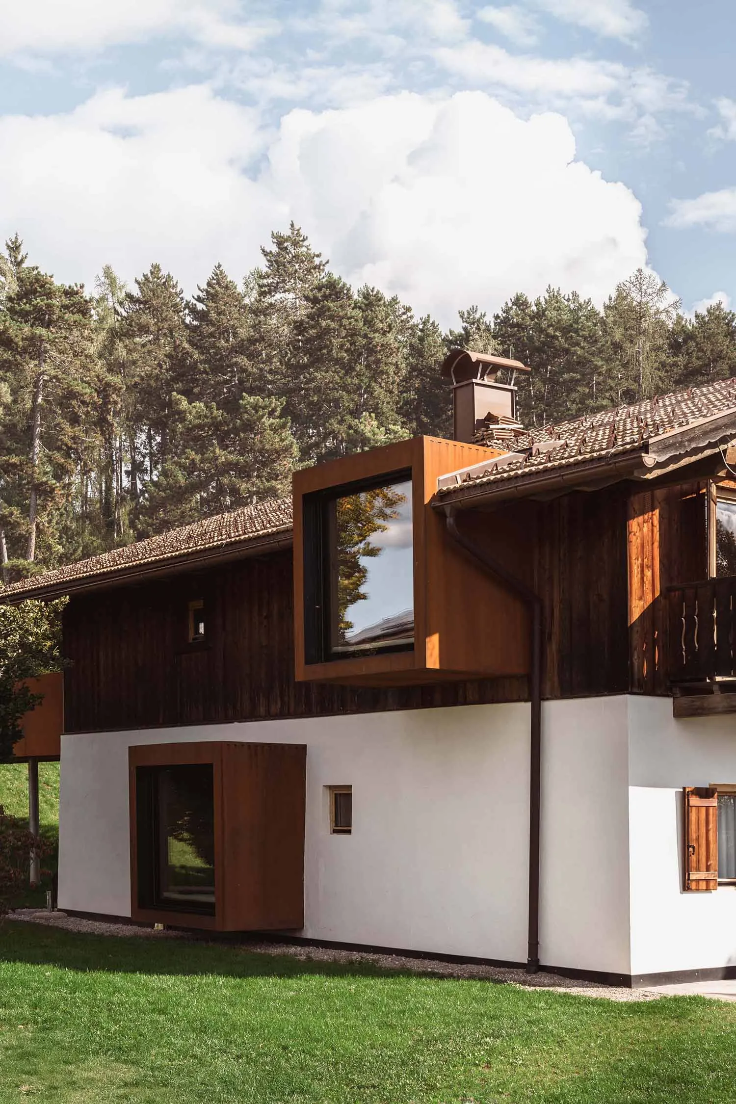 Exterior view of a contemporary mountain house featuring a large cantilevered Corten steel box with black-framed windows, set against a dense pine forest under a blue sky.