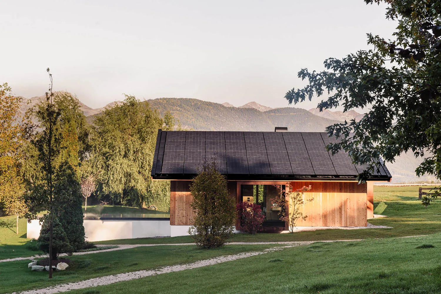 Modern wooden cabin with black solar panel roof, surrounded by green lawn, trees and distant mountains at golden hour.