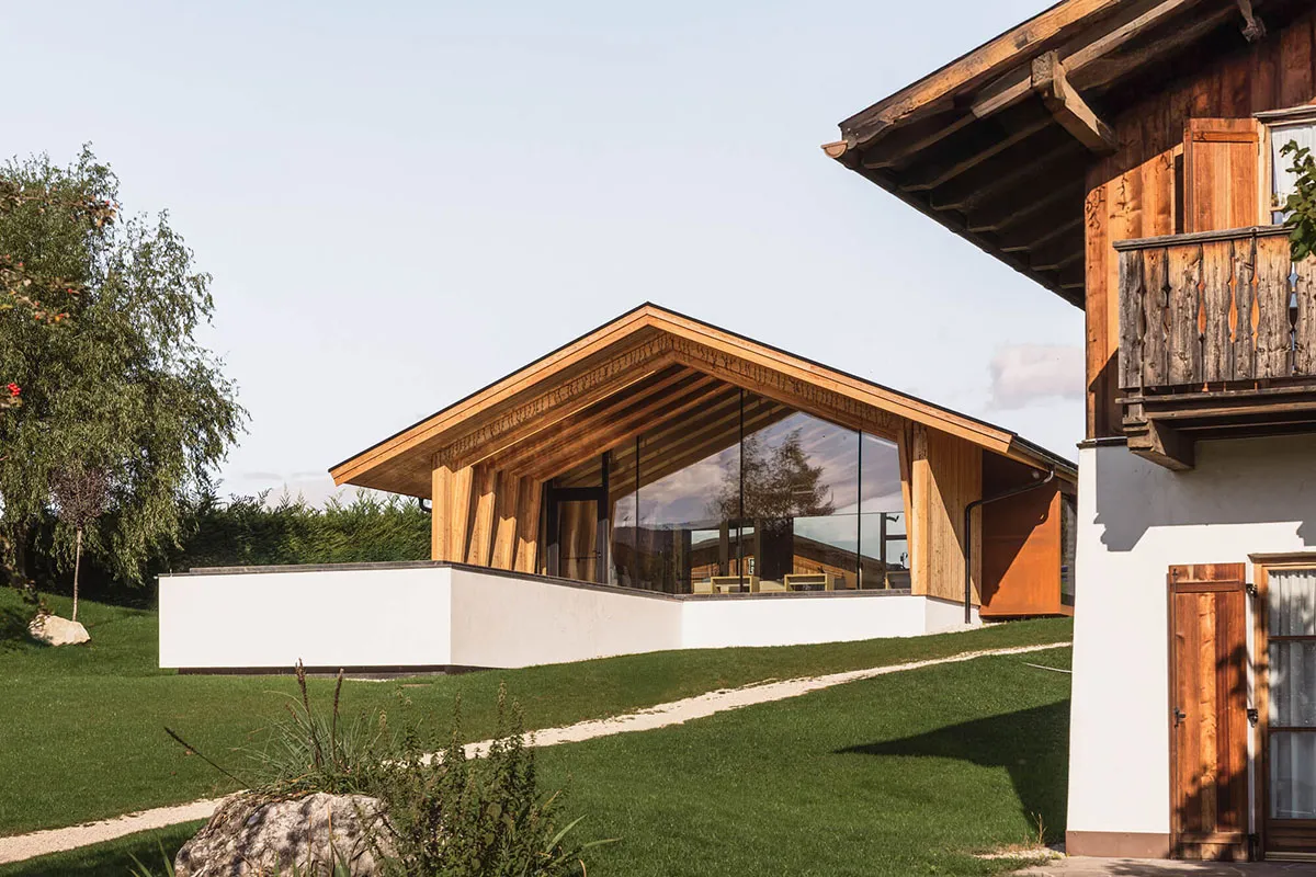 Modern alpine mountain house featuring large glass walls, exposed wooden beams, white concrete base and traditional wooden balcony under a clear sky.