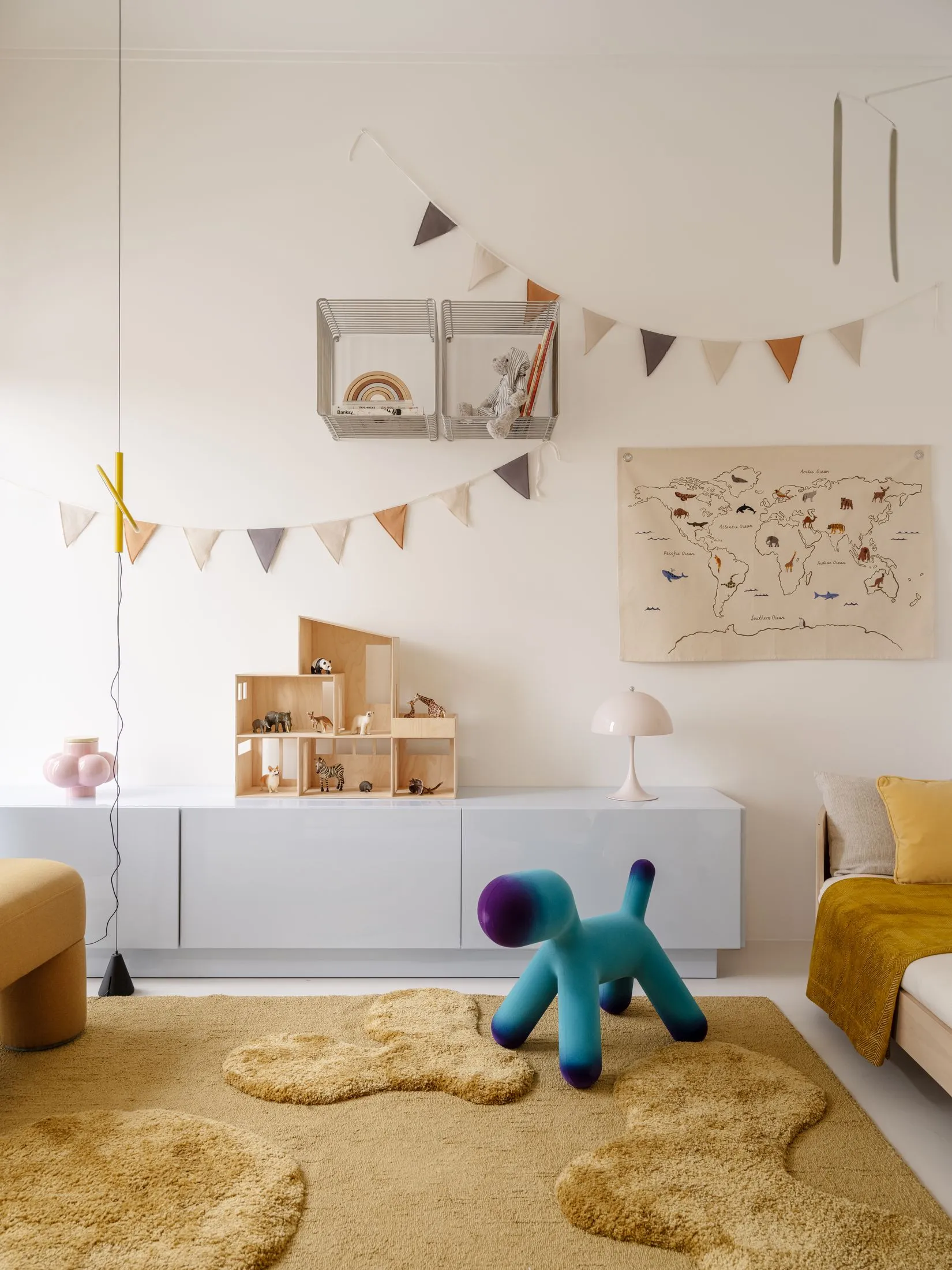 Bright and airy children's playroom with white storage, wooden dollhouse, animal figurines, cloud-shaped beige felt rugs, a blue-purple gradient dog stool, and a world map on the wall.