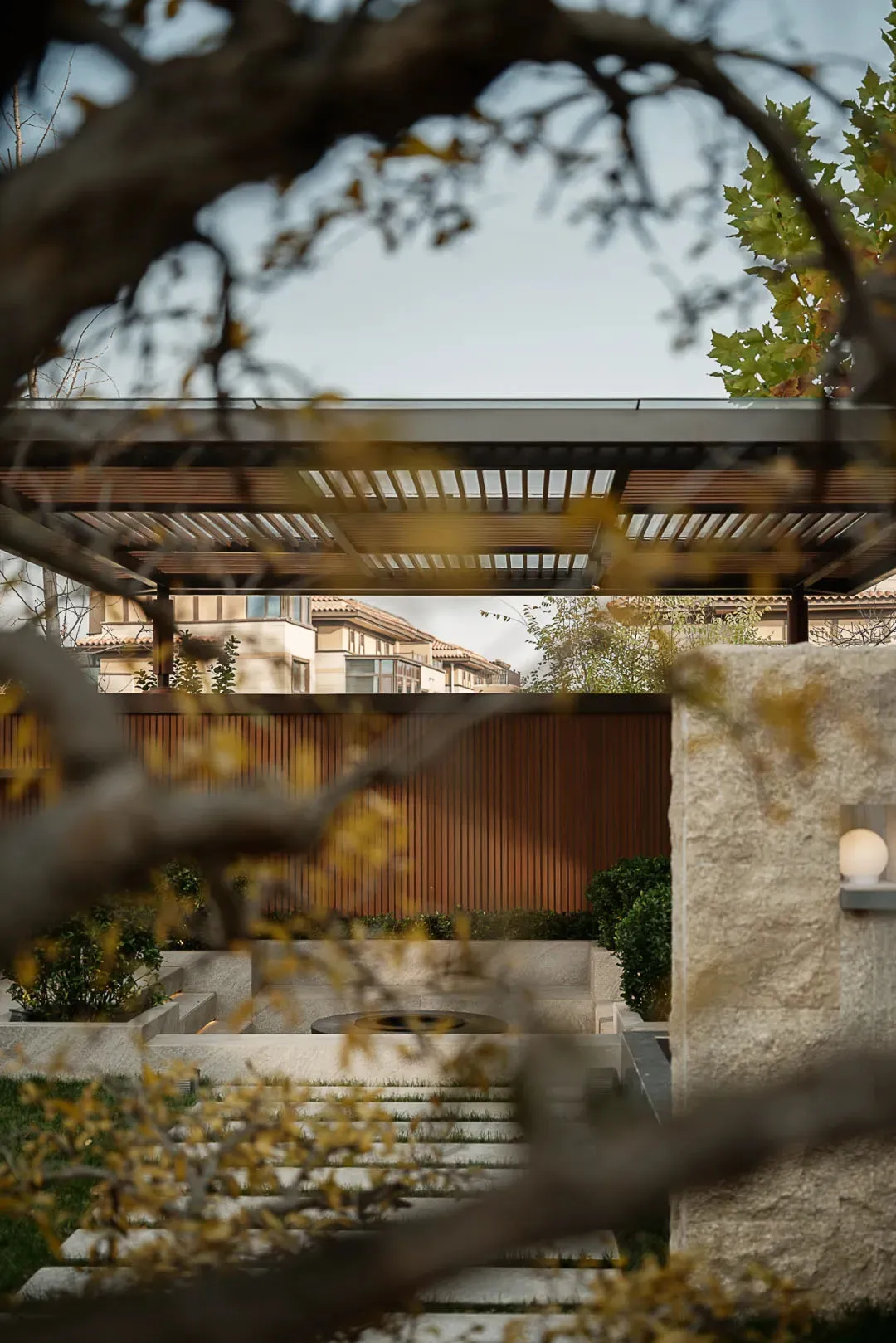 Modern sunken courtyard garden featuring layered travertine steps, a circular fire pit, vertical wood slat wall, and overhead timber pergola with integrated lighting.