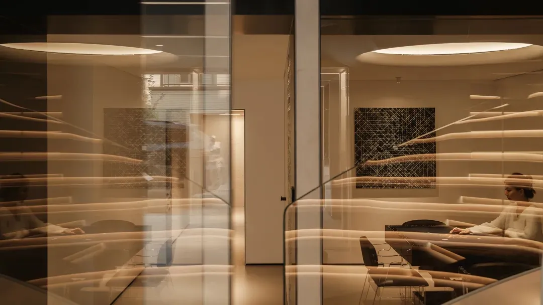 Modern dining room viewed through multiple layers of curved glass partitions with warm ambient lighting, abstract black artwork, and a white sculpture in the background.