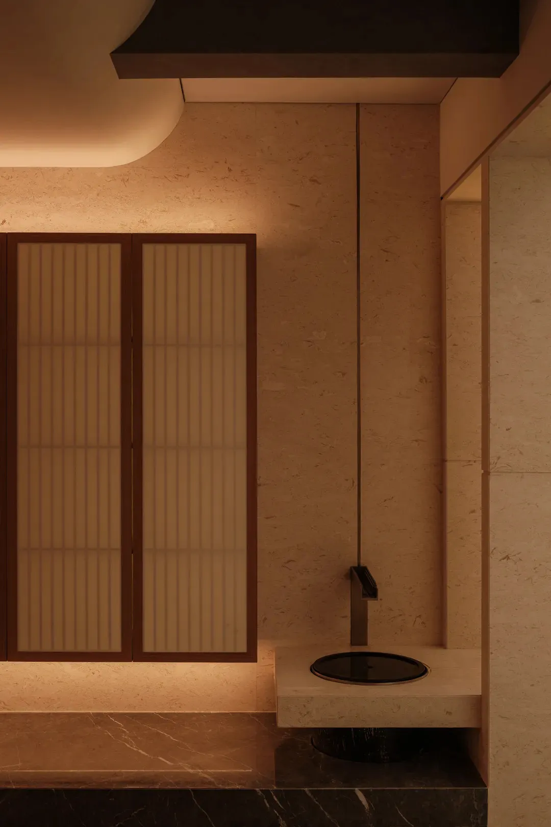 Modern minimalist powder room featuring a wall-mounted black circular sink, matte black faucet, travertine stone walls and floor, dark marble base, and backlit wooden shoji-style cabinet doors.