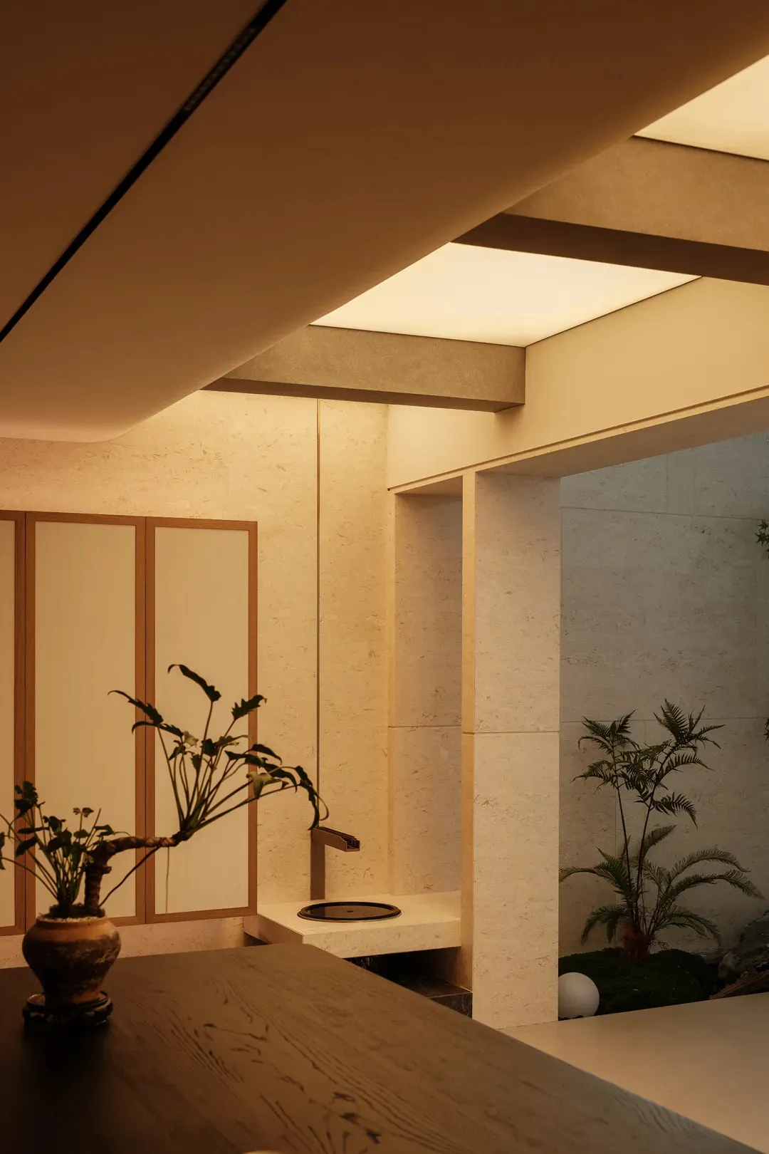 Modern Japanese minimalist interior featuring a small indoor courtyard with travertine stone walls, wooden bonsai planter, and integrated water feature under soft warm lighting.