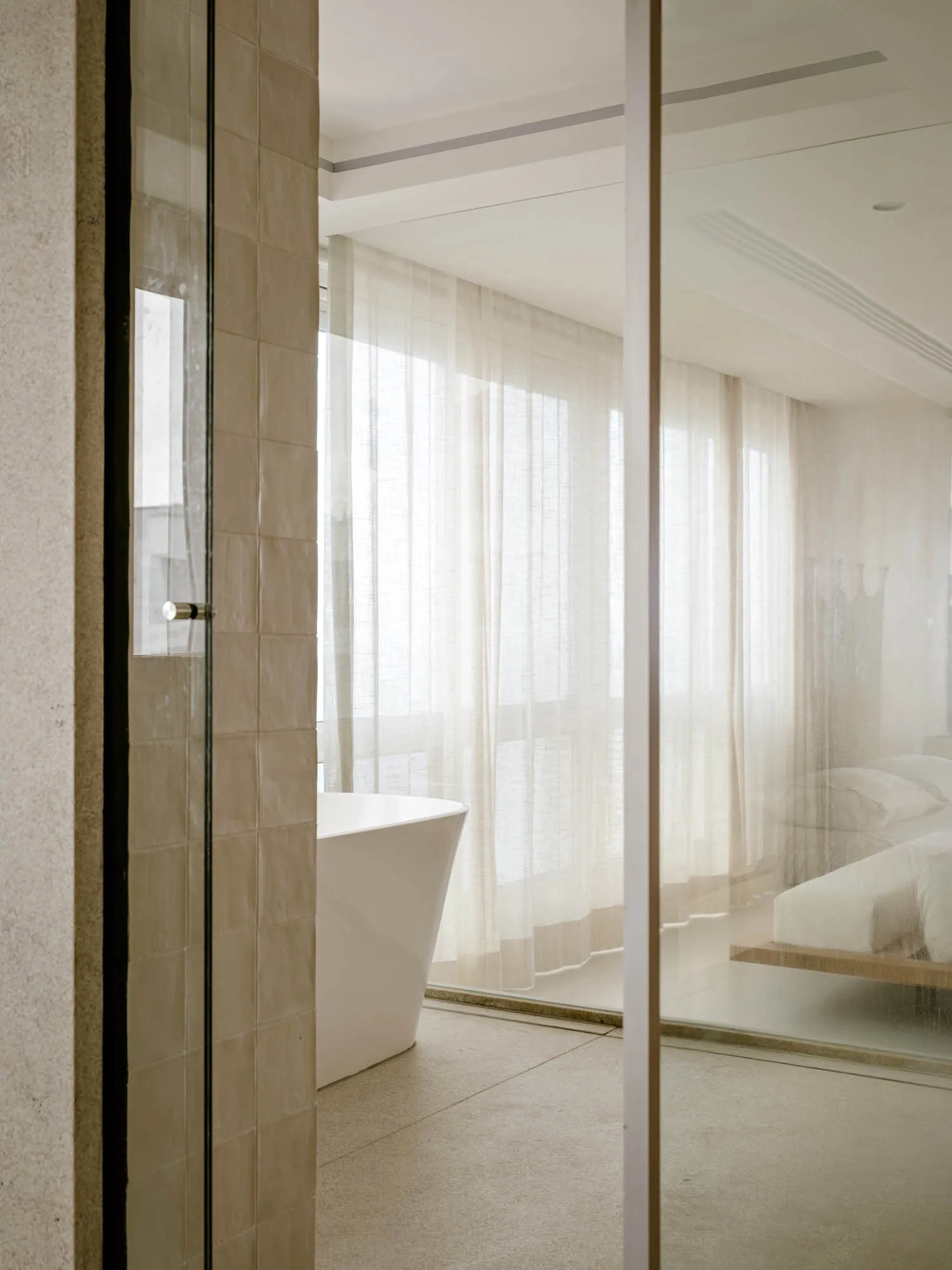 Modern minimalist bathroom interior with beige travertine tiled walls, white freestanding bathtub, sheer white curtains, and a floating bed visible through glass partition.