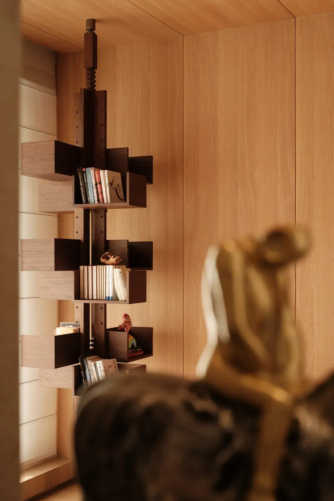 Modern sculptural walnut bookshelf with staggered cantilevered shelves filled with books, displayed in a warm wood-paneled reading corner with soft ambient lighting.