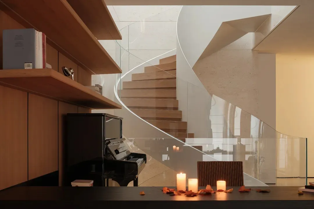 Modern interior featuring a sweeping curved oak staircase, black grand piano, built-in wooden bookshelves, and lit candles on a dark table with autumn leaves.