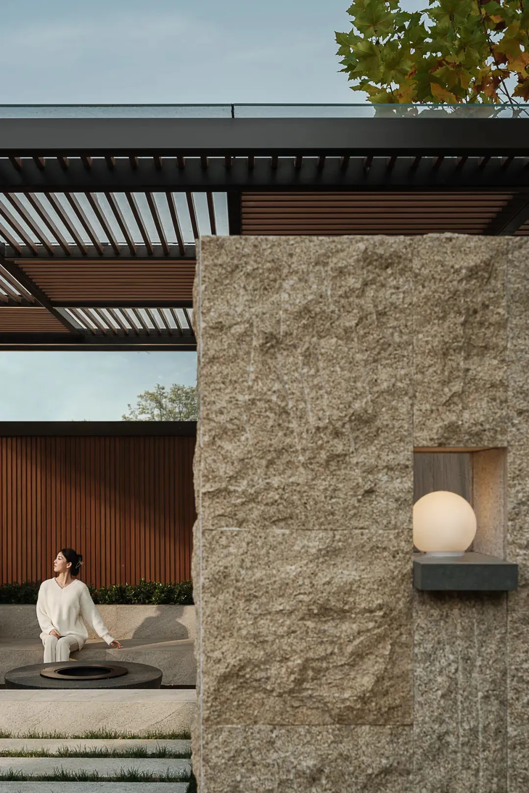 Modern outdoor courtyard featuring a large travertine stone wall with a recessed glowing sphere lamp, wooden pergola, circular fire pit, and a woman in white sitting on stone steps.
