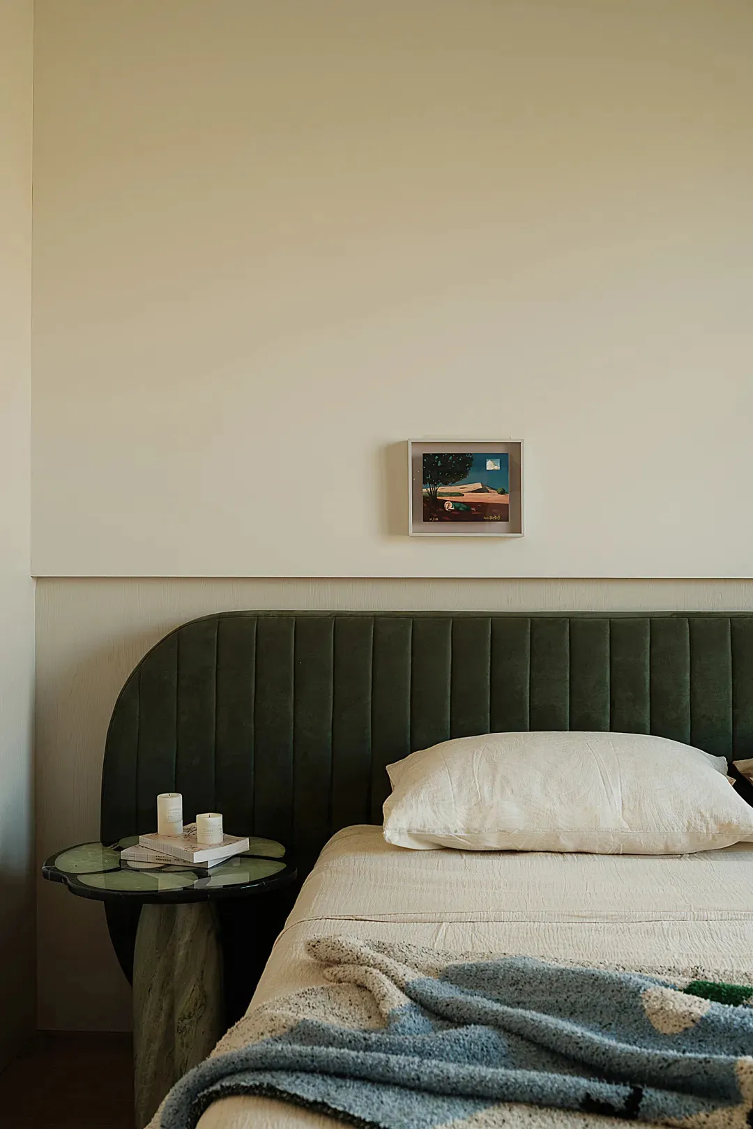 Modern bedroom with curved dark green velvet headboard, white linen bedding, blue textured throw blanket, and a unique green marble side table with candles and books against a two-tone wall with framed art.
