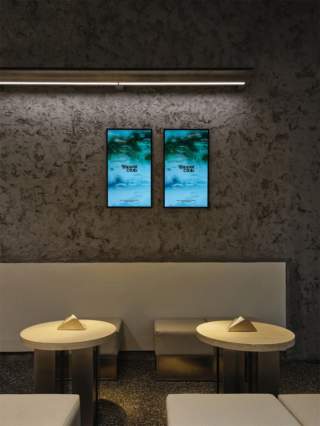 Modern lounge area featuring two round wooden tables, upholstered benches, and two framed Ripple Club posters mounted on a textured concrete wall under a linear LED light bar.