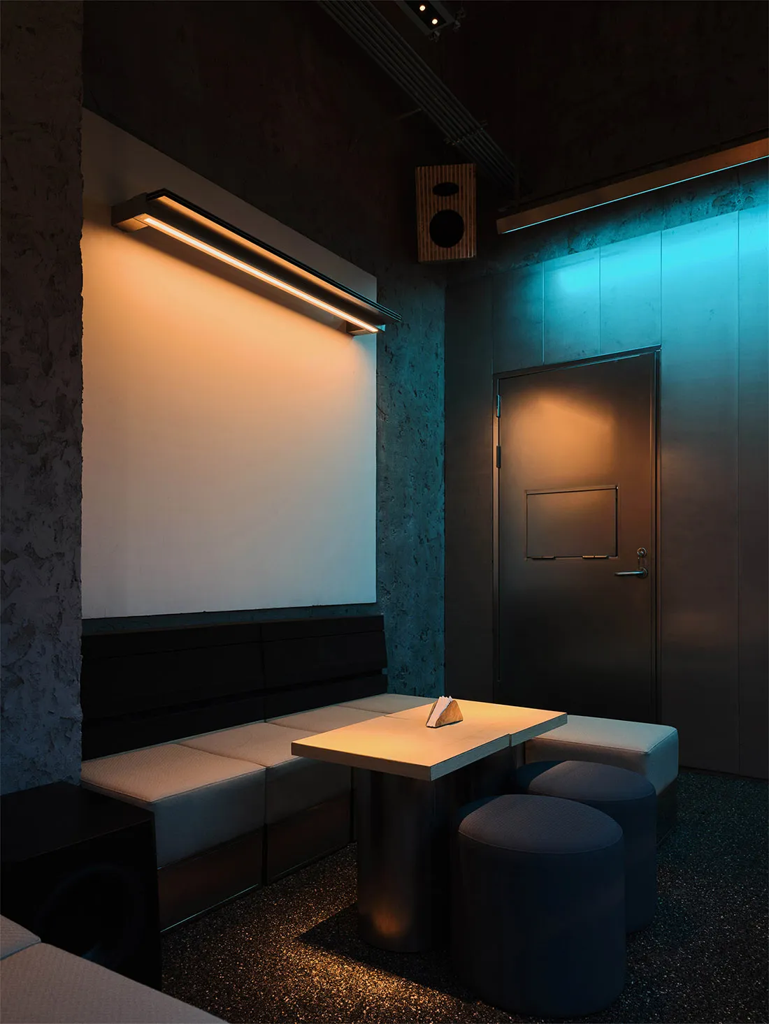Modern industrial private dining area featuring a built-in upholstered booth, concrete walls, a rectangular dining table, round stools, and dramatic dual-tone lighting with warm white and cool blue hues.
