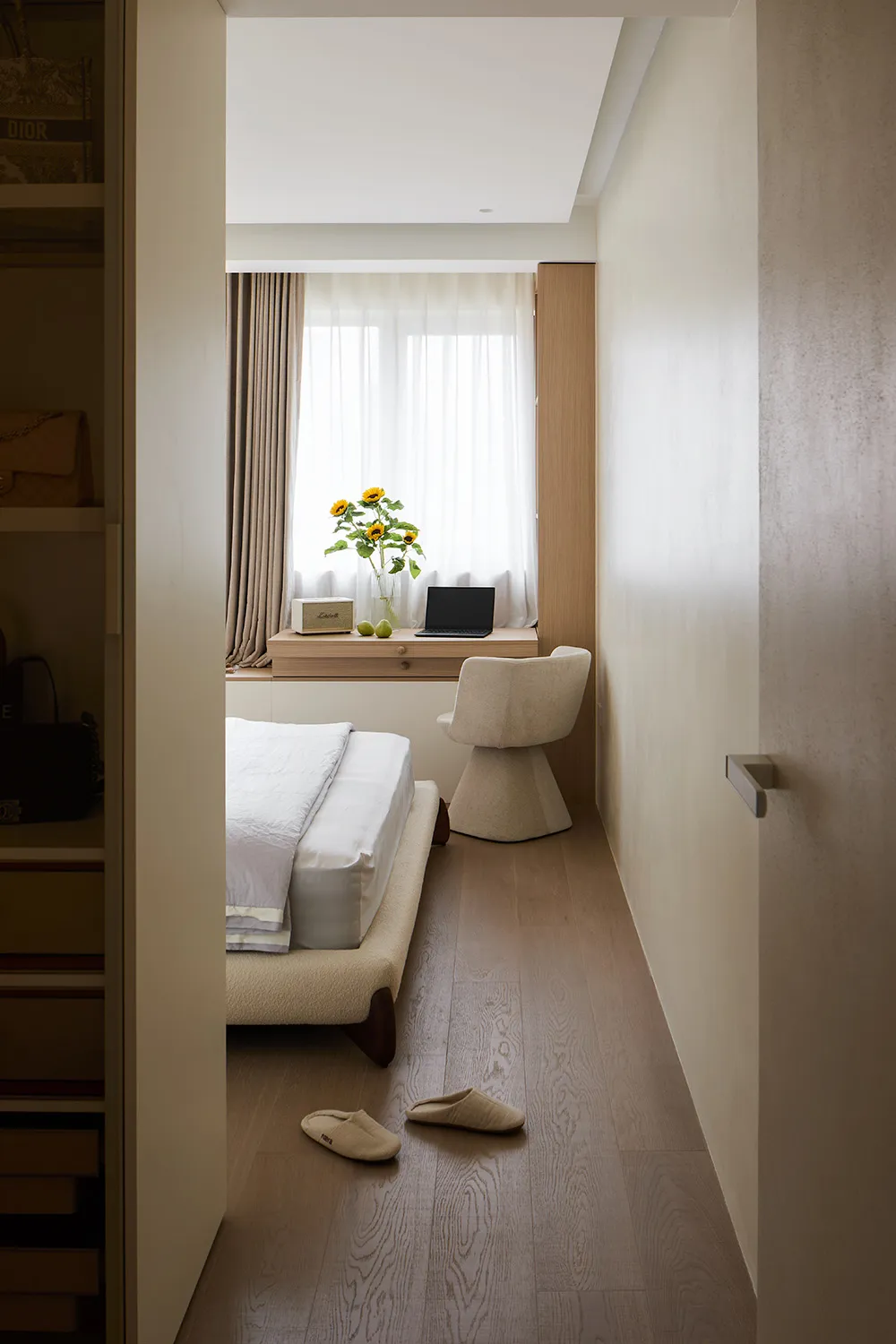View into a minimalist bedroom featuring a low-profile bed with white linens, a boucle swivel chair at a built-in window desk, sheer curtains, and light wood flooring.