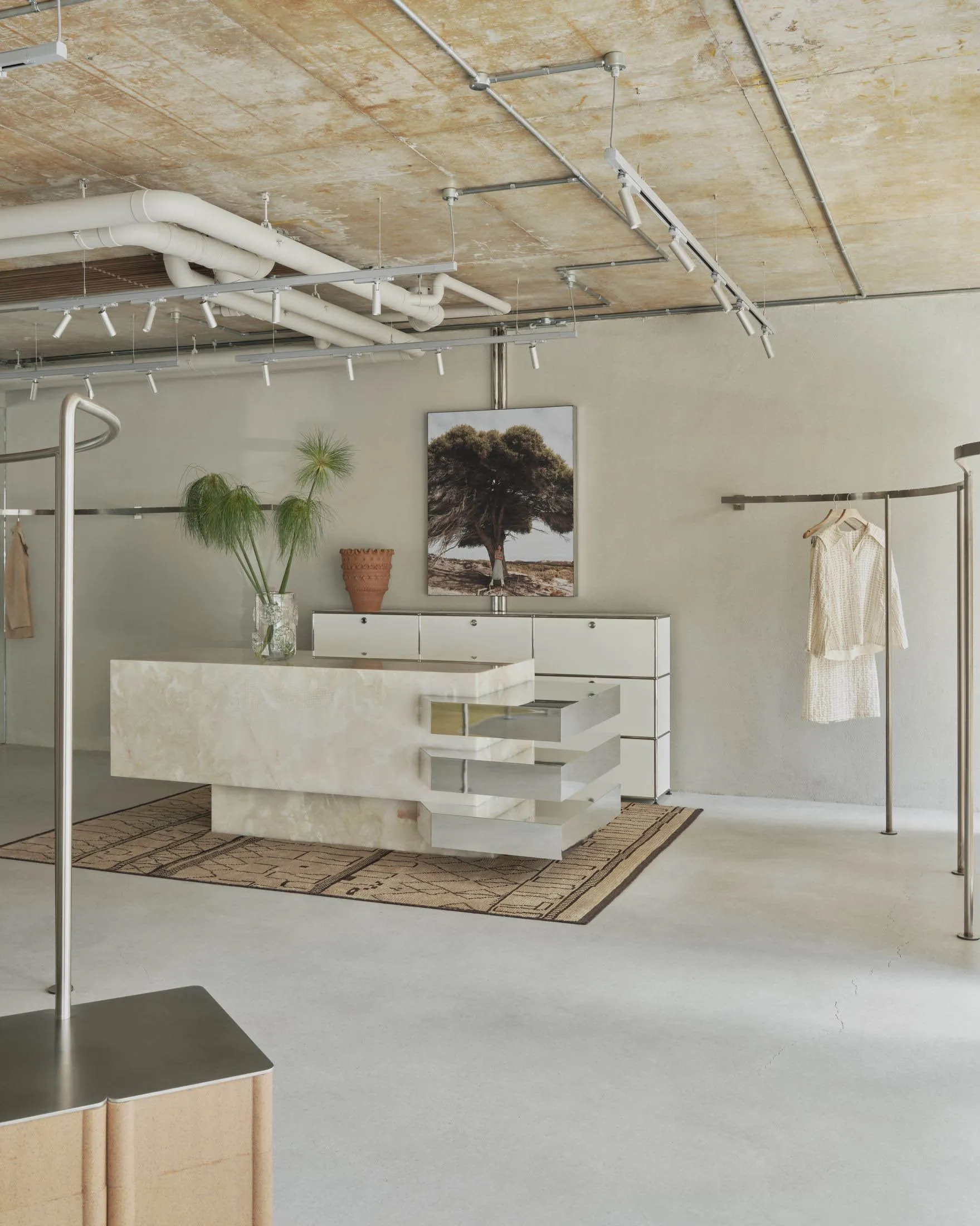 Modern minimalist boutique interior featuring a large travertine stone central island, white modular cabinetry, industrial track lighting, and clothing racks under an exposed concrete ceiling.