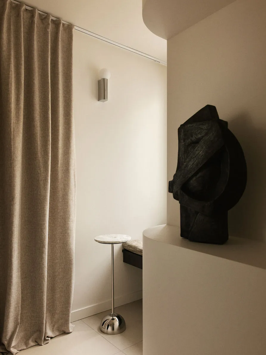 Modern minimalist entryway corner featuring a dark abstract stone sculpture on a curved white pedestal, a slender travertine-top side table with chrome base, beige linen curtain, and cylindrical wall sconce.