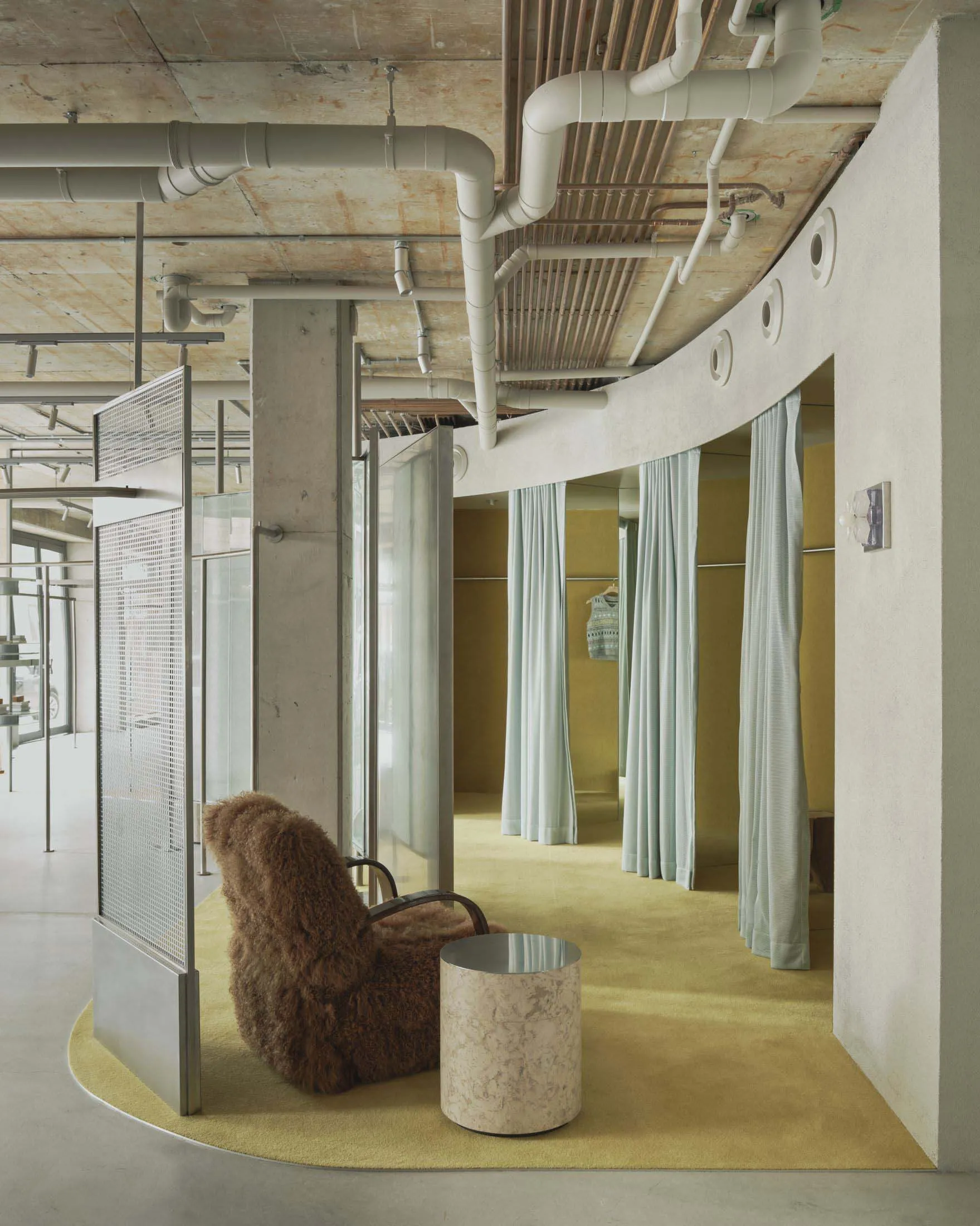 Modern fitting room area with curved concrete wall, exposed ceiling pipes, chartreuse carpet, brown sheepskin-covered armchair and marble side table