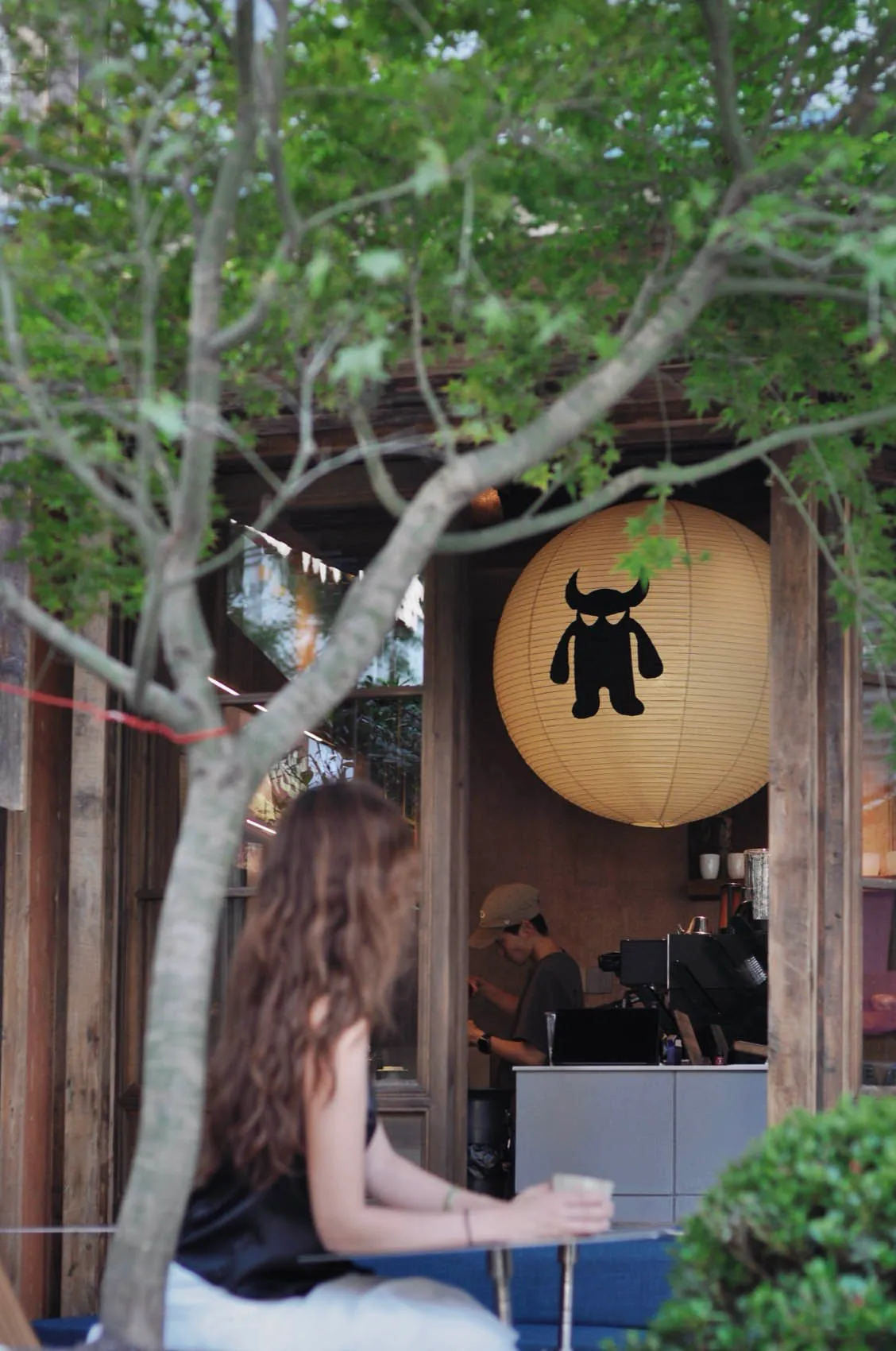Woman sitting at an outdoor cafe table under a tree, with a large yellow paper lantern featuring a black monster logo hanging at the wooden entrance of the shop.