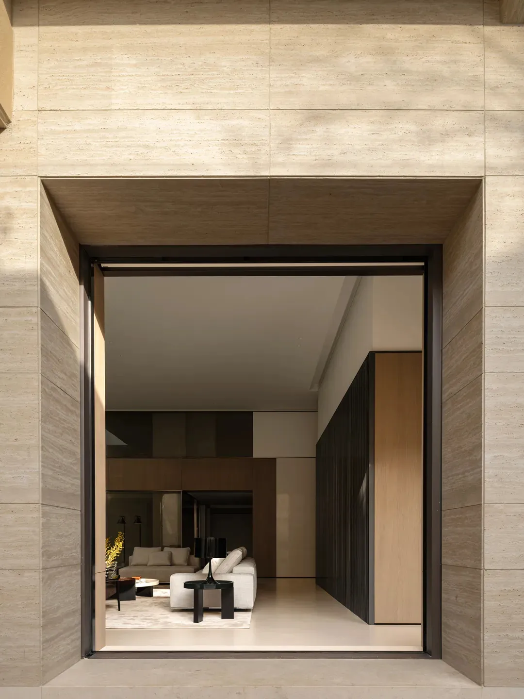 View through a large travertine stone doorway into a modern minimalist living room with neutral furnishings and dark wood accents