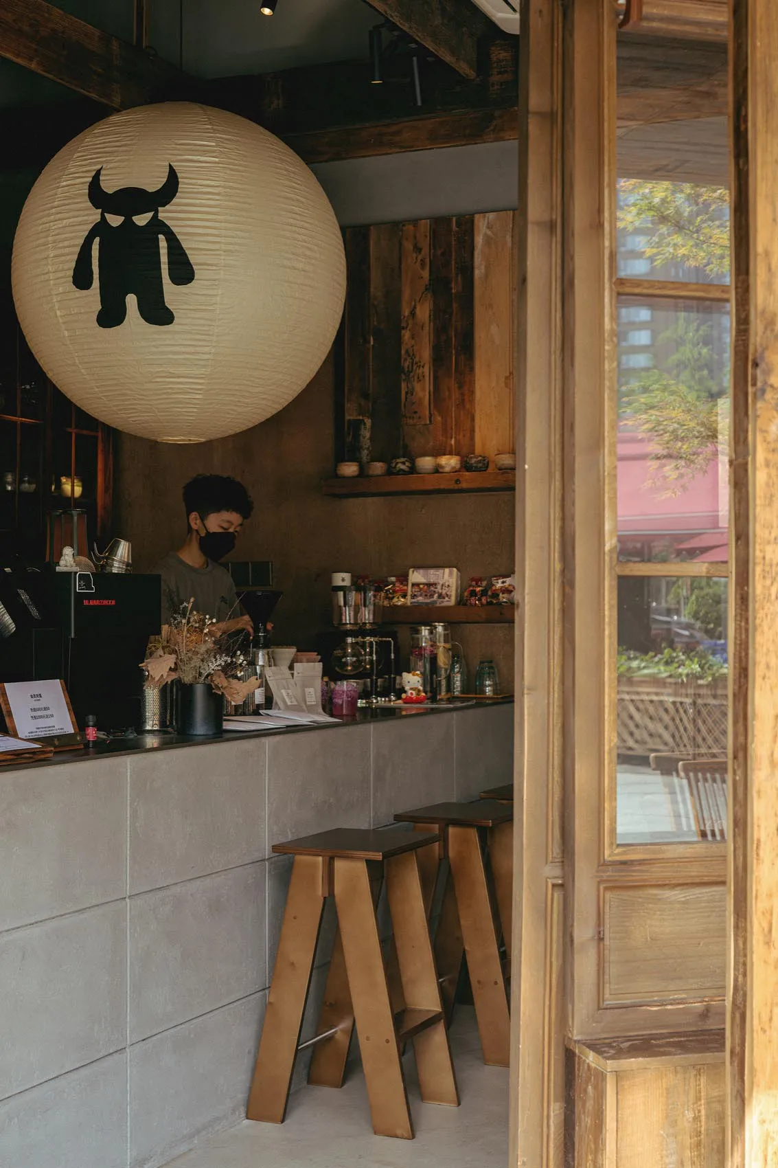 View of a minimalist cafe counter with concrete block base, wooden stools, a barista working, and a large white paper lantern with a black monster graphic hanging from the ceiling.