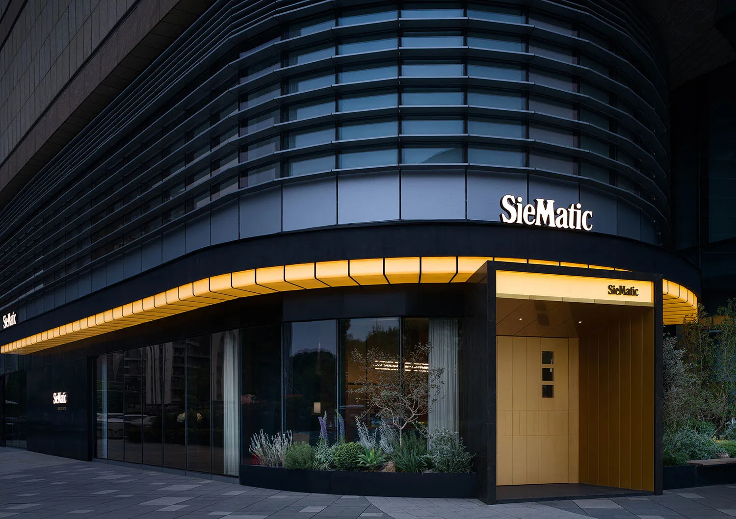 Night view of a modern luxury kitchen showroom with a curved dark glass facade, warm yellow LED lighting along the canopy, and landscaped planters at the entrance.