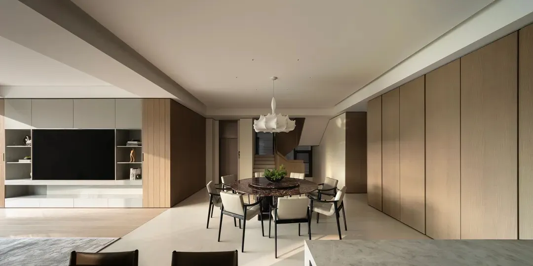 Modern minimalist dining room featuring a large round dark marble table surrounded by six light-colored dining chairs, under a white sculptural pendant lamp, with floor-to-ceiling walnut wood paneling and built-in media console in the background.