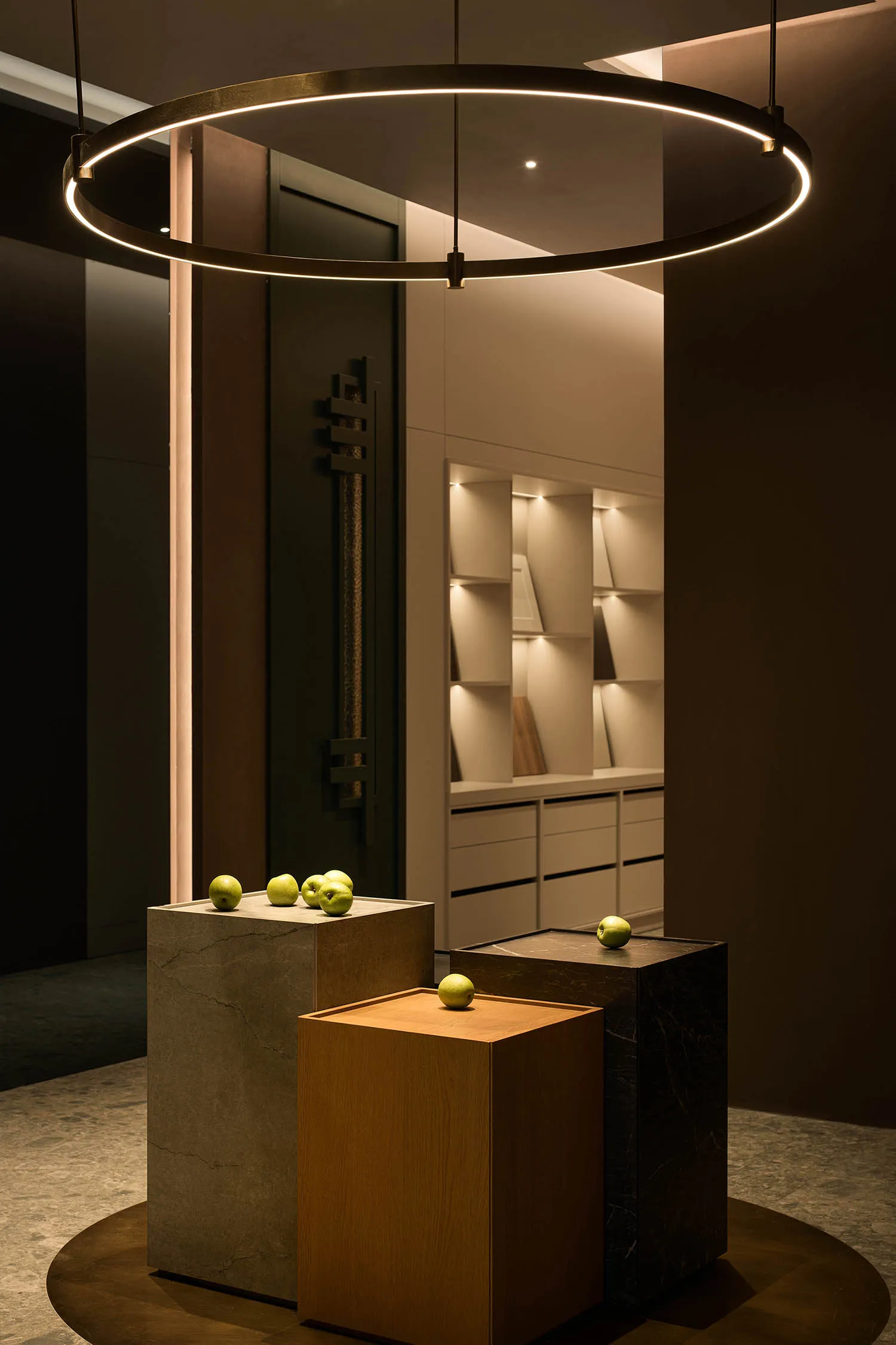 Modern minimalist entryway featuring a large circular LED pendant lamp, three display pedestals in concrete, light oak, and black marble, each topped with green apples, with built-in illuminated shelving in the background.