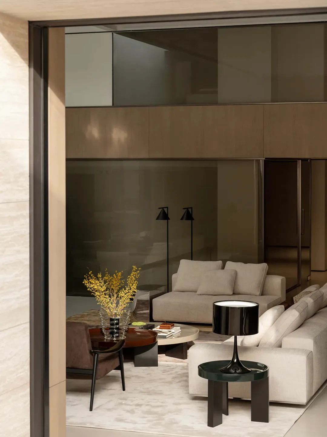 Modern luxury living room featuring a large beige sectional sofa, matching armchair, round coffee tables, black floor lamps, and warm wood paneling in the background.