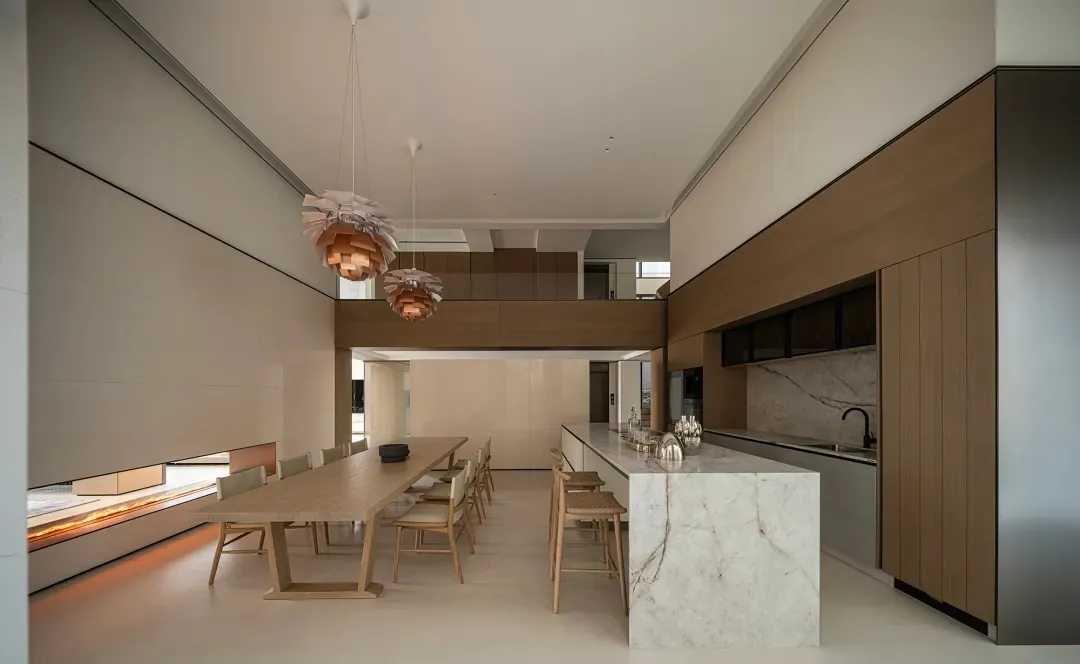 Modern double-height open-plan kitchen and dining area featuring light oak dining table, marble kitchen island, and two large wooden pendant lights hanging from high ceiling.