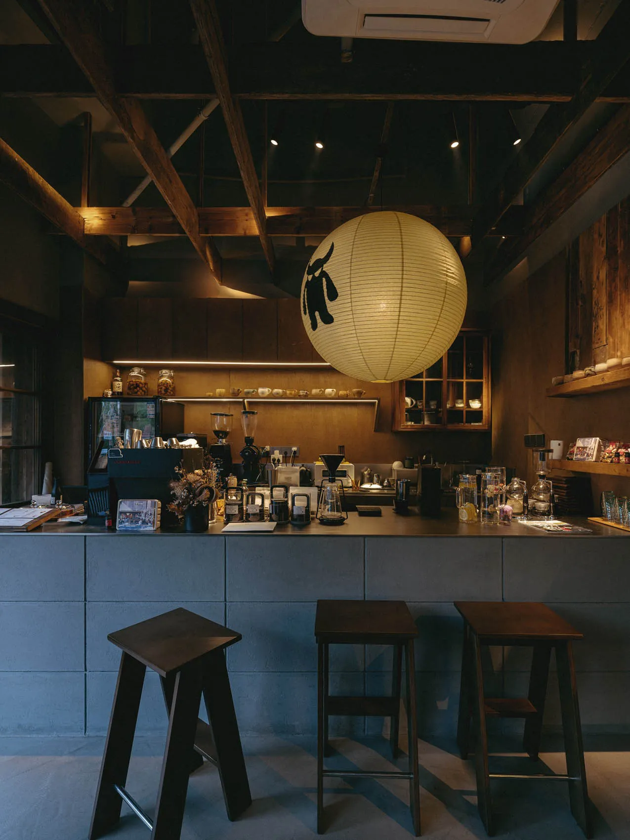 Interior view of a specialty coffee shop with dark wood stools, concrete counter, wooden ceiling beams, and a large illuminated Japanese paper lantern with a bull logo.