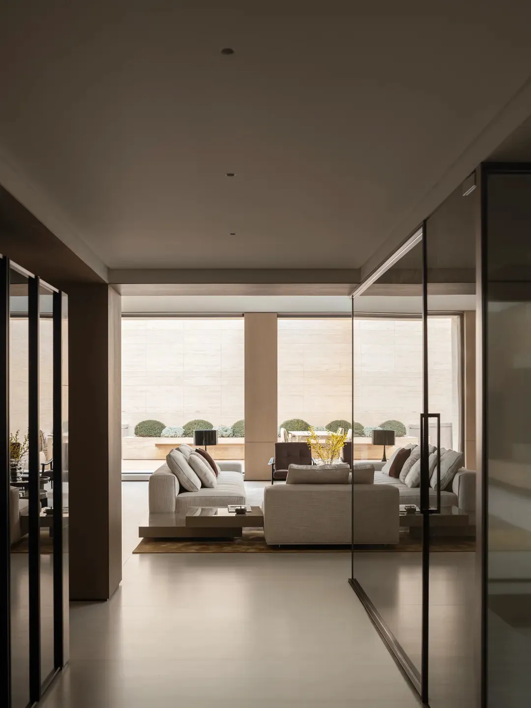 Modern minimalist living room featuring light travertine walls and floor, neutral fabric sofas, and large glass doors opening to a landscaped garden with trimmed shrubs.