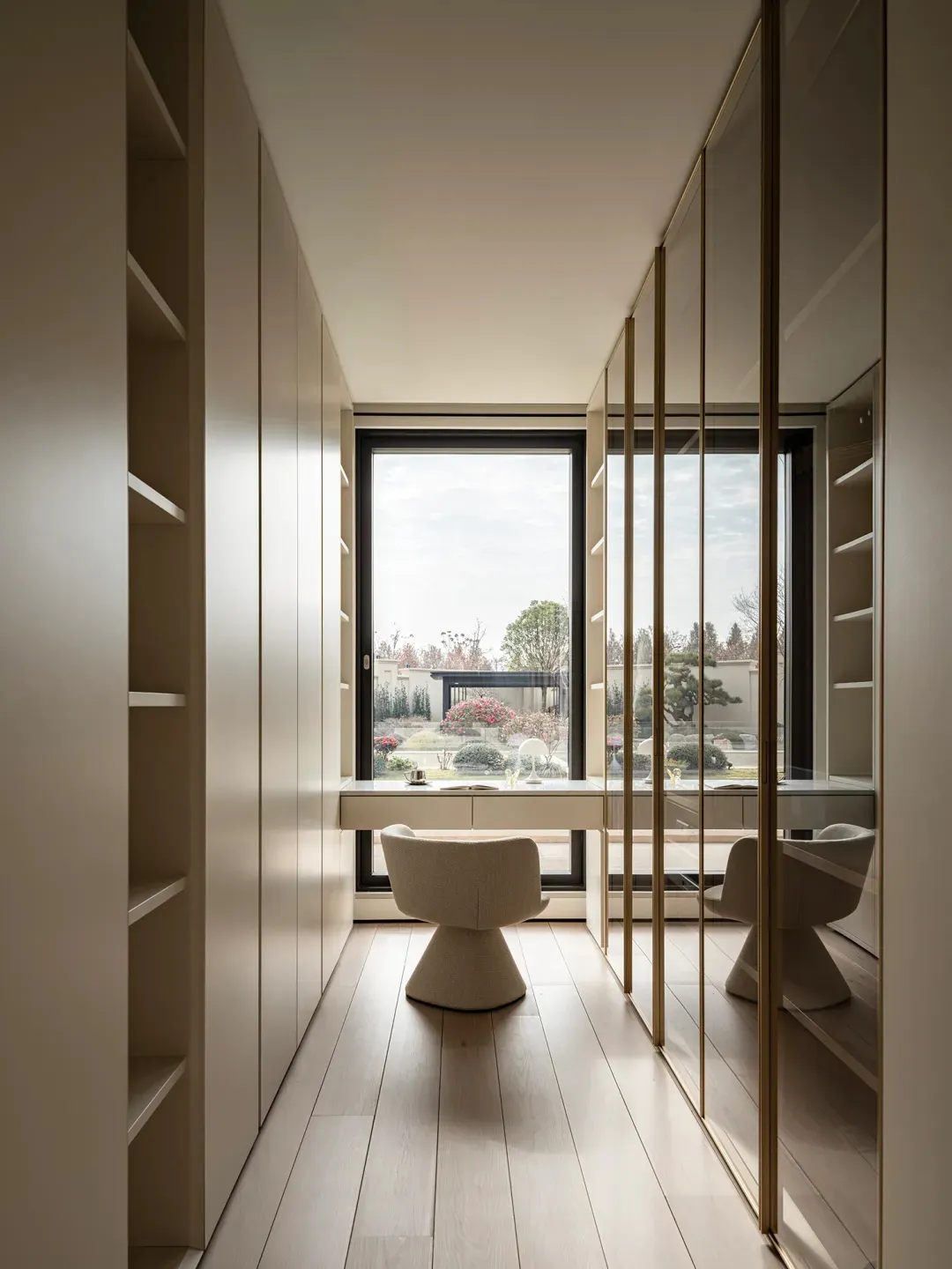 Modern narrow home office featuring built-in beige lacquer cabinetry, a large picture window overlooking a landscaped garden, a white bouclé swivel chair, and brass-framed glass doors.