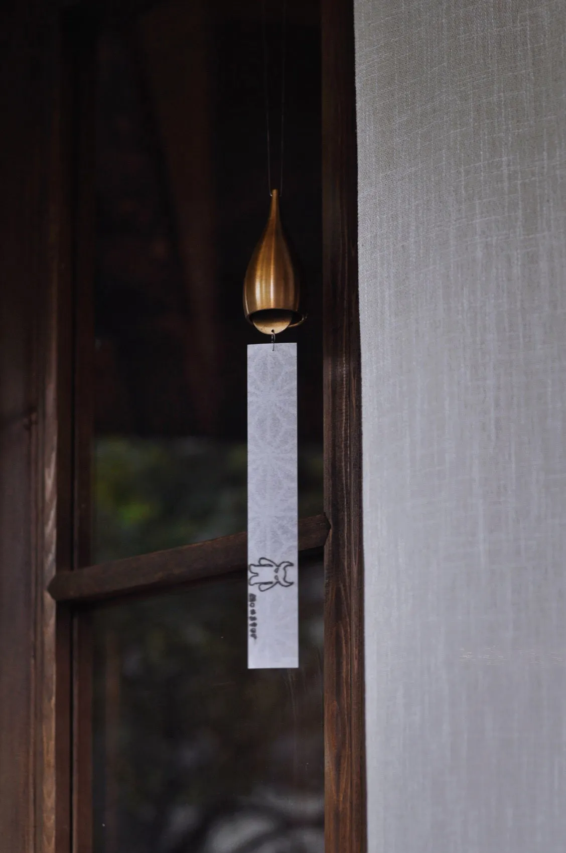 A brass teardrop-shaped wind chime with a white patterned paper strip hanging in front of a dark wooden Japanese-style window with blurred greenery outside.