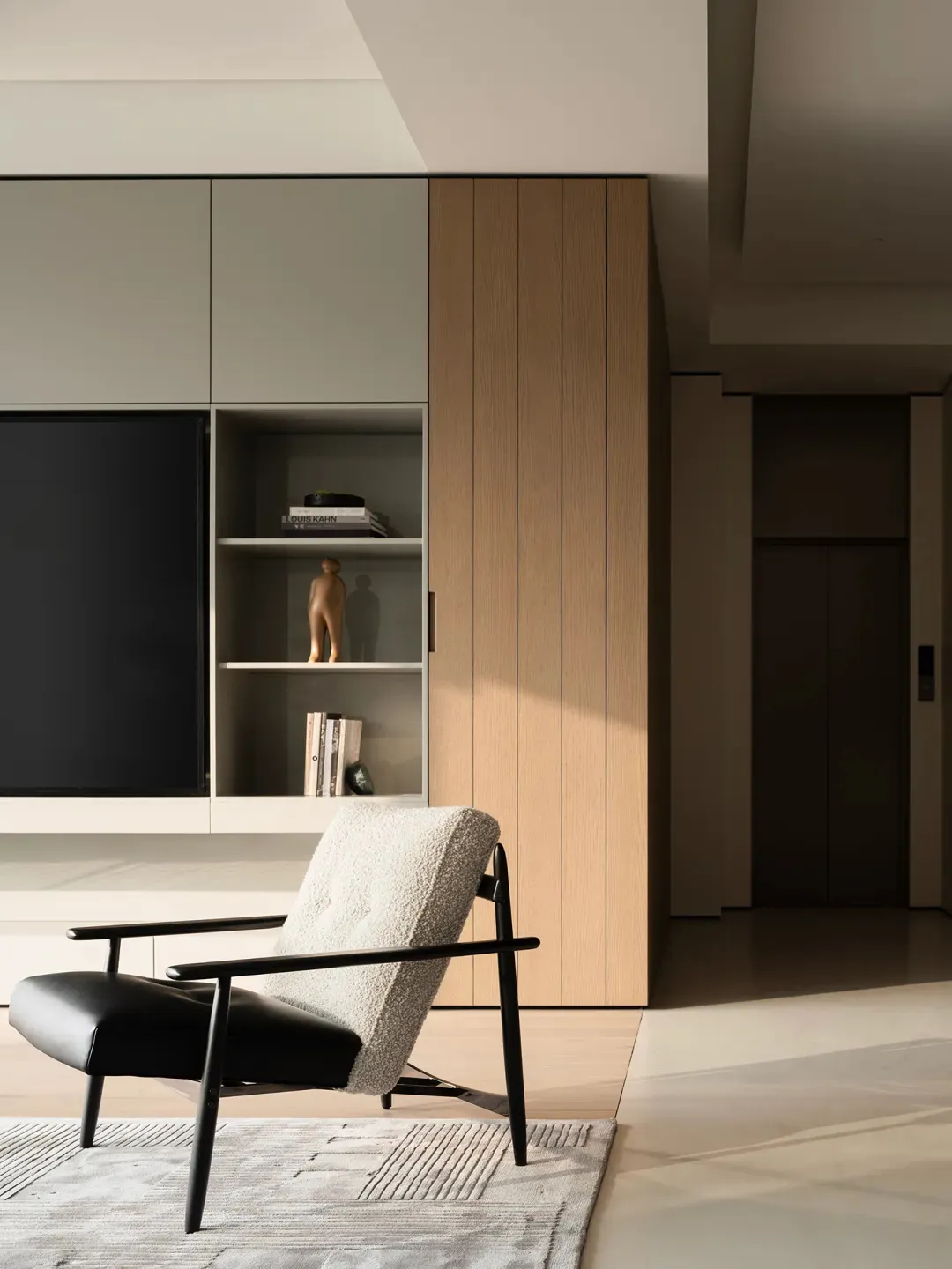 Modern living room featuring a black and white boucle armchair with black metal frame on a textured rug, positioned in front of built-in light wood and off-white cabinetry with open shelving and a large television.