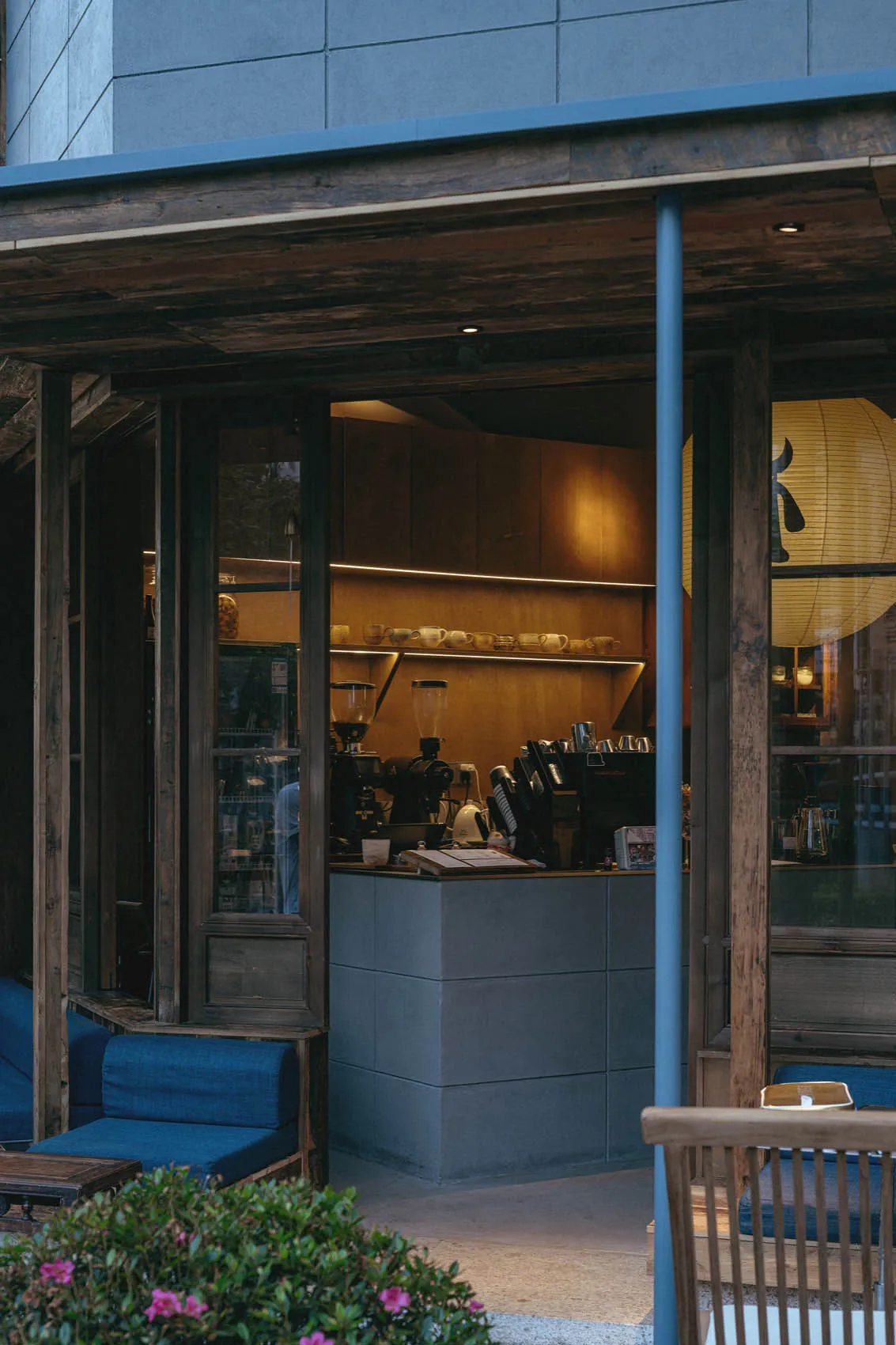 View from an outdoor wooden terrace into a warmly lit Japanese-style cafe interior featuring concrete counter, reclaimed wood beams, and deep blue upholstered seating.