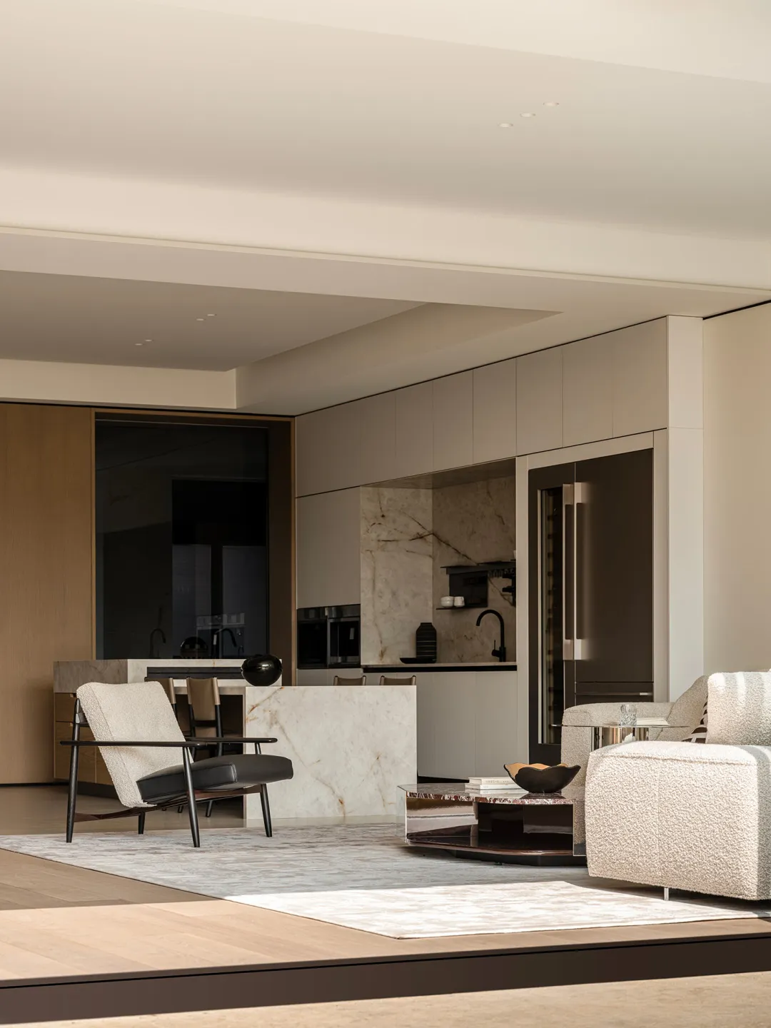 Modern minimalist living room featuring a white marble kitchen island, boucle sofa, black leather and wood armchair, and integrated stainless steel appliances.