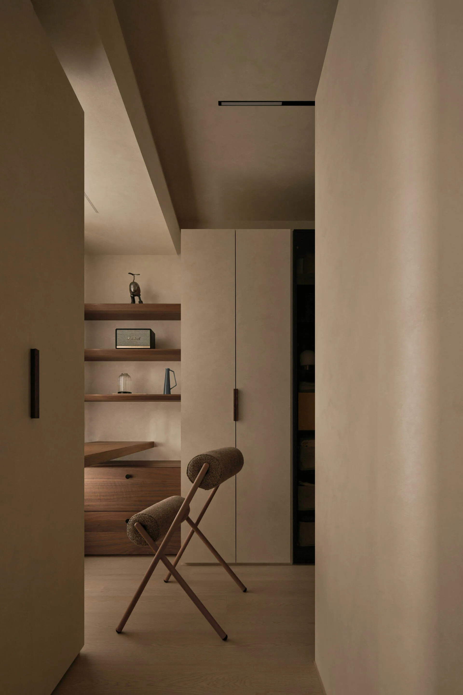 Minimalist dressing room featuring walnut floating shelves, built-in drawers, a tall beige wardrobe, and a wooden X-frame stool with bouclé upholstery.