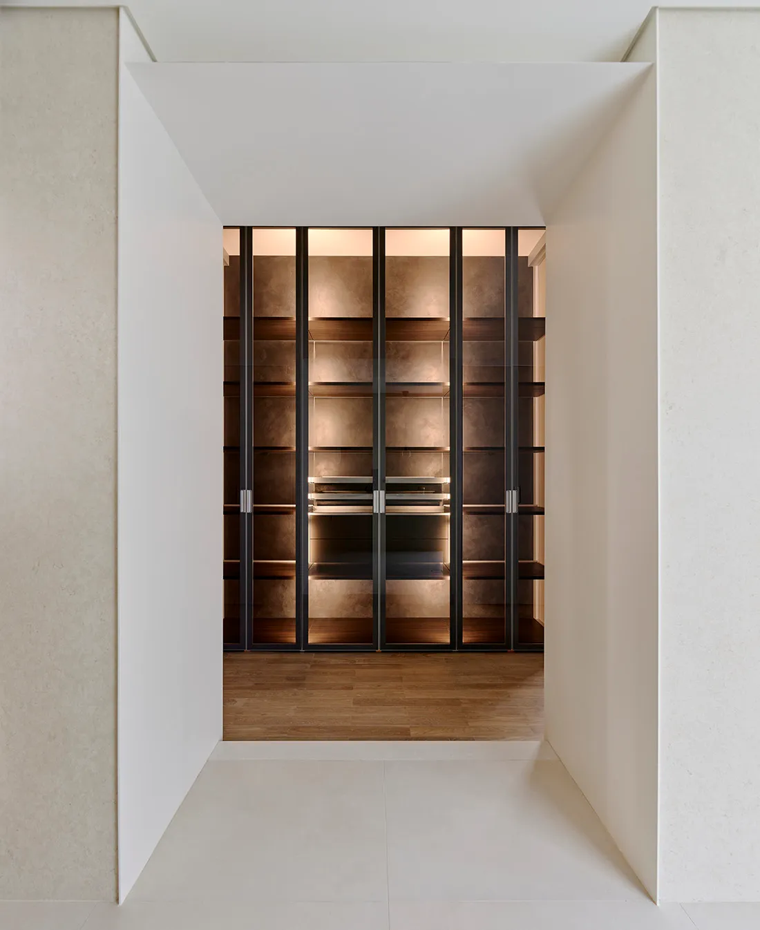 Modern minimalist walk-in closet featuring dark metal framed glass doors and backlit walnut wood open shelving with warm lighting.