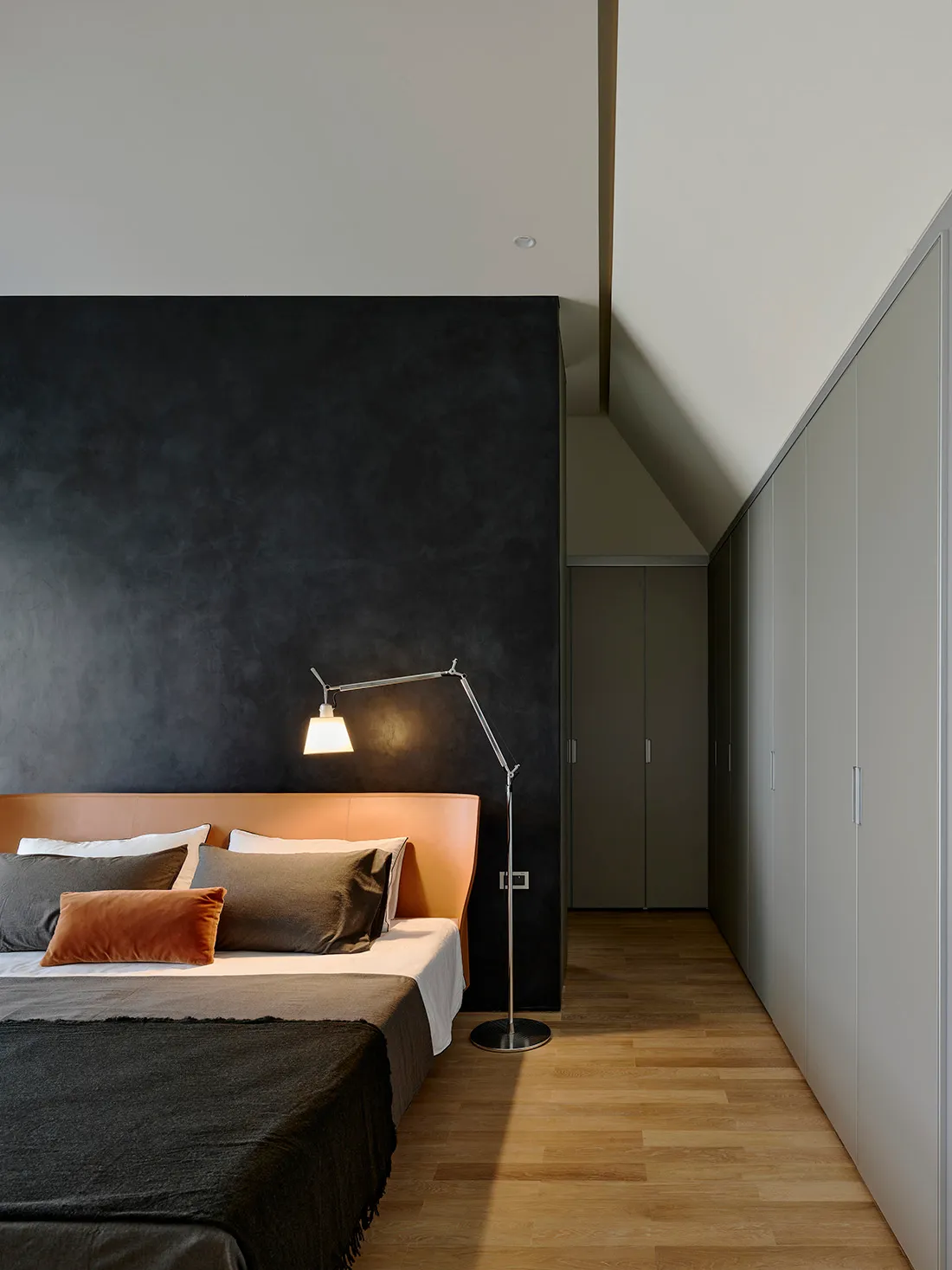 Modern minimalist bedroom with a large dark charcoal textured feature wall, tan leather upholstered headboard, layered grey and rust pillows, and a chrome articulated floor lamp.
