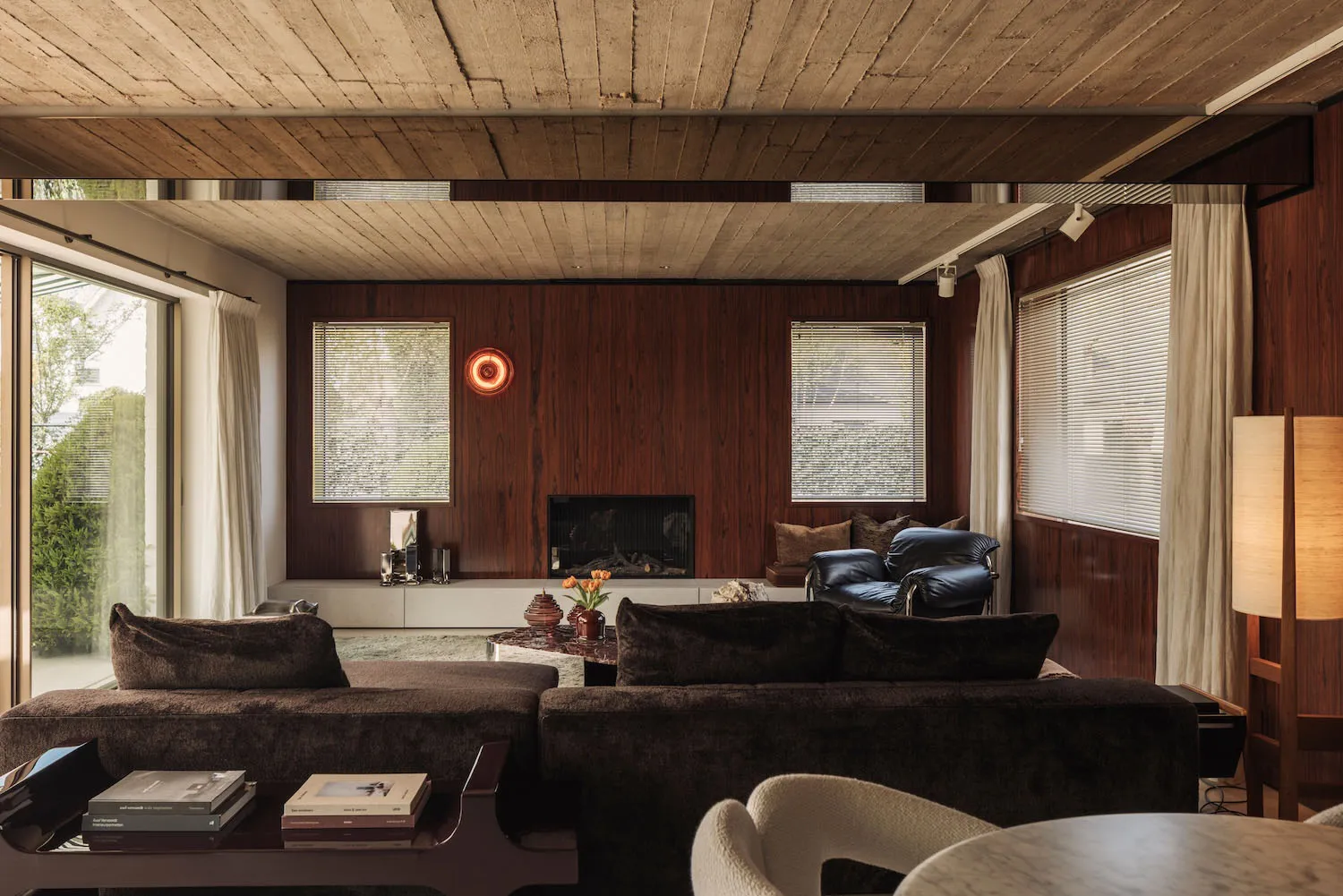 Mid-century modern living room with dark walnut wood paneling, brown velvet sectional sofa, leather lounge chairs, and a wooden ceiling.