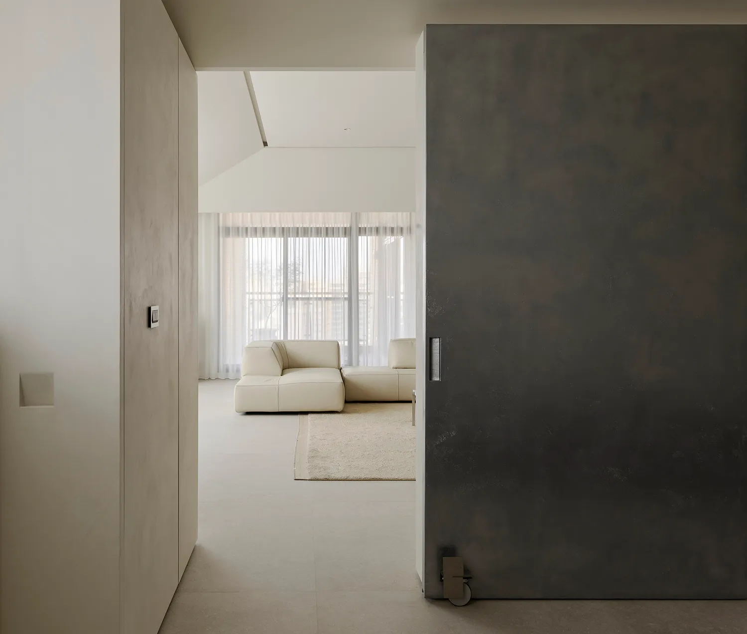 View from a concrete hallway into a bright minimalist living room featuring off-white modular sofa, large windows with sheer curtains, and micro-cement walls.