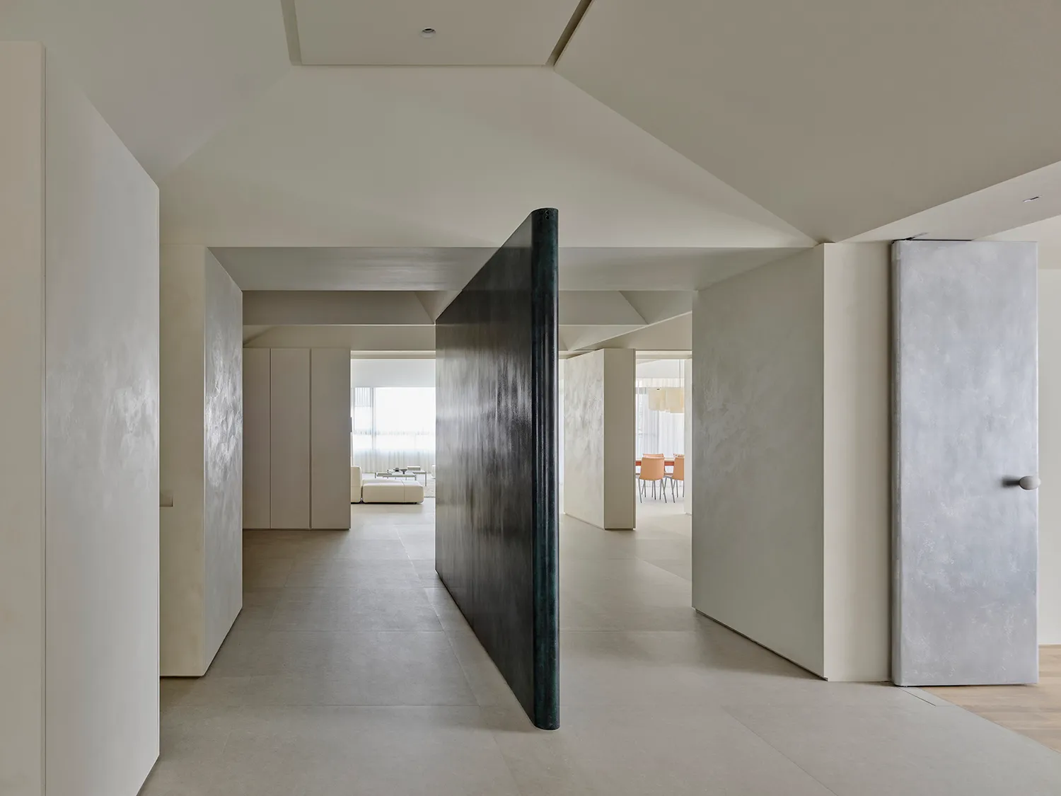 Modern minimalist hallway featuring light micro-cement walls and floors, a tall dark metal pivot door, and views into adjacent living and dining areas.