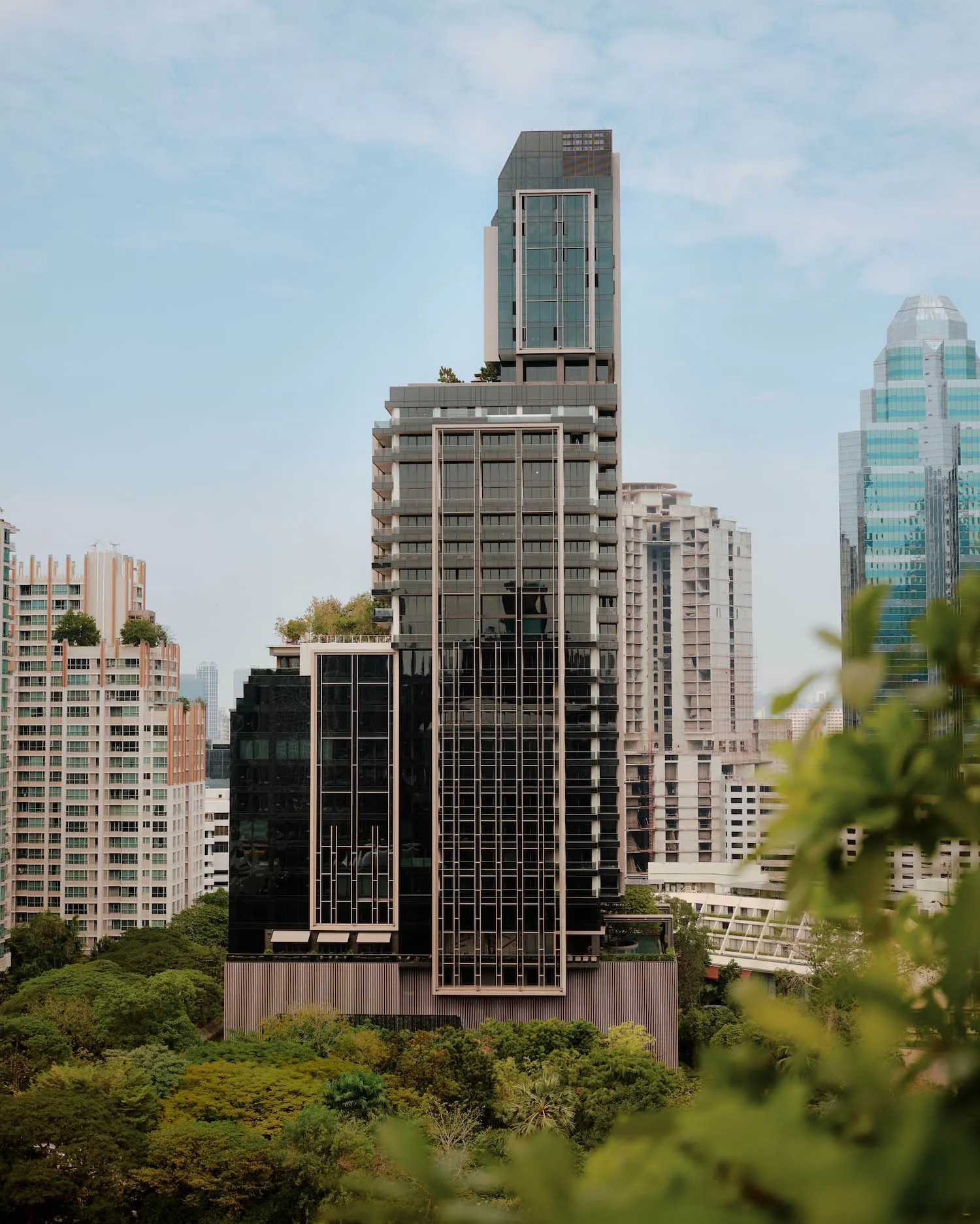 Tall modern residential skyscraper with dark glass facade and vertical lines, surrounded by dense green trees and other city buildings under a blue sky.