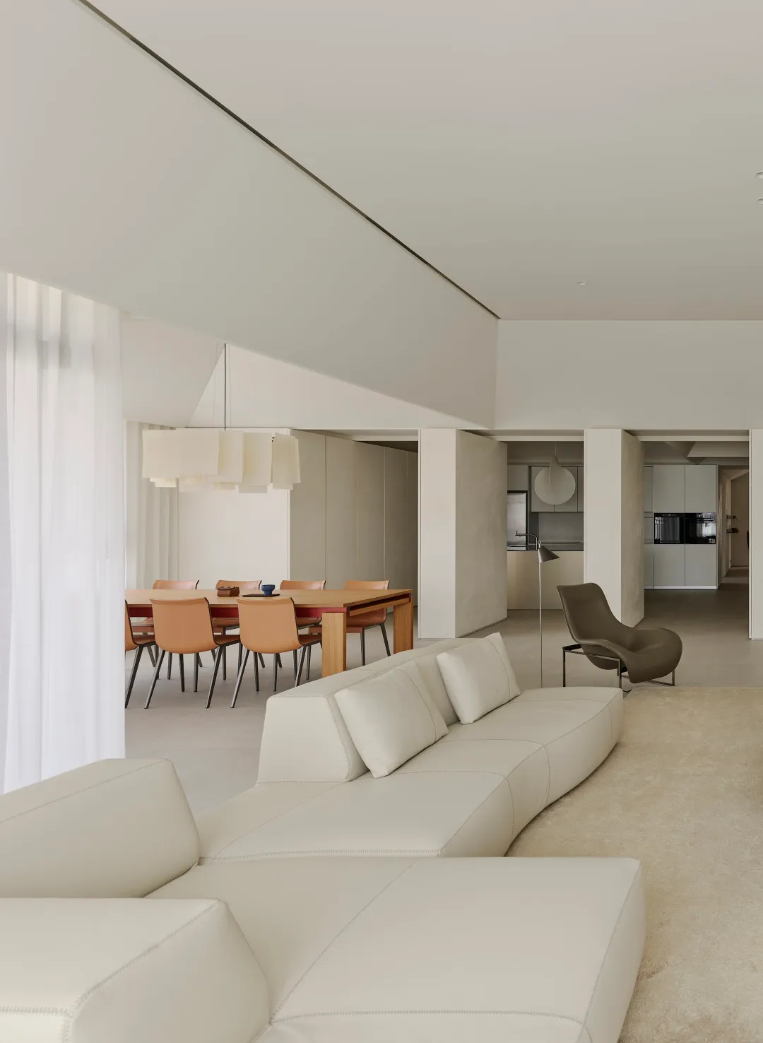 Modern minimalist open-plan living room featuring a long curved white leather sofa, warm oak dining table with leather chairs, and a sculptural olive lounge chair under high ceilings.