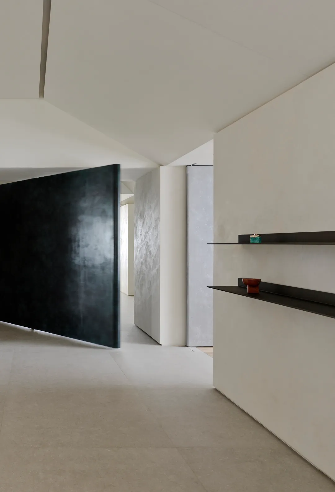 Modern minimalist hallway featuring a large black pivoting door, light micro-cement walls, floating black shelves with decorative objects, and seamless concrete flooring.