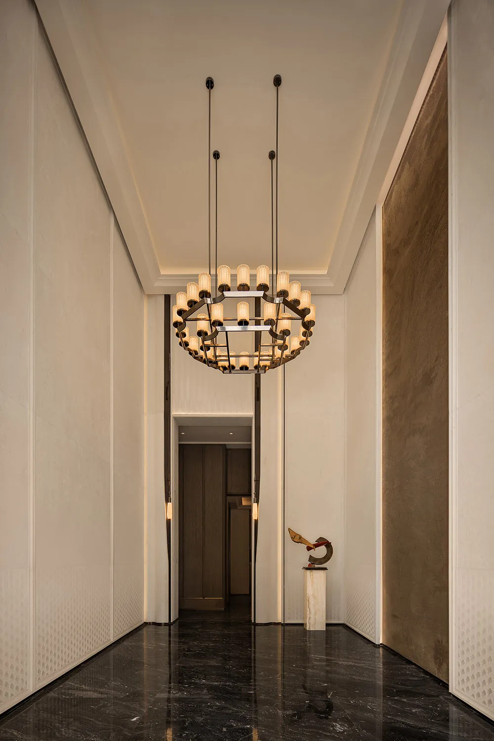Modern luxury hallway with black marble floor, white textured walls, large contemporary chandelier, and abstract sculpture on a marble pedestal.