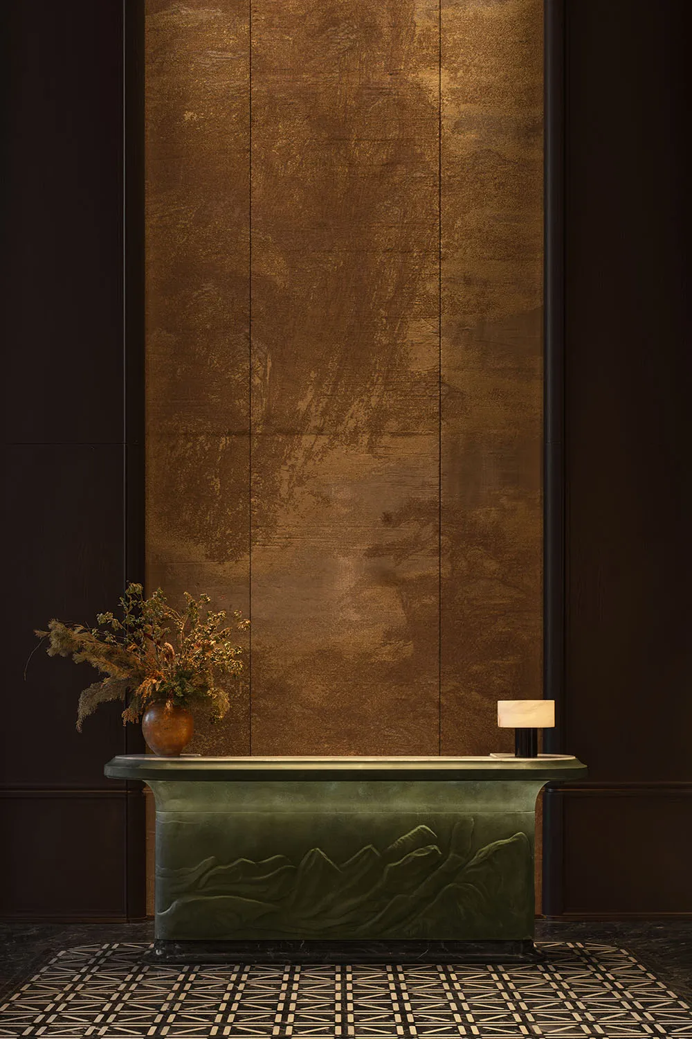 Luxury hotel reception desk in verdigris green with mountain relief carving, placed against a textured gold leaf wall with dried floral arrangement and marble lamp.