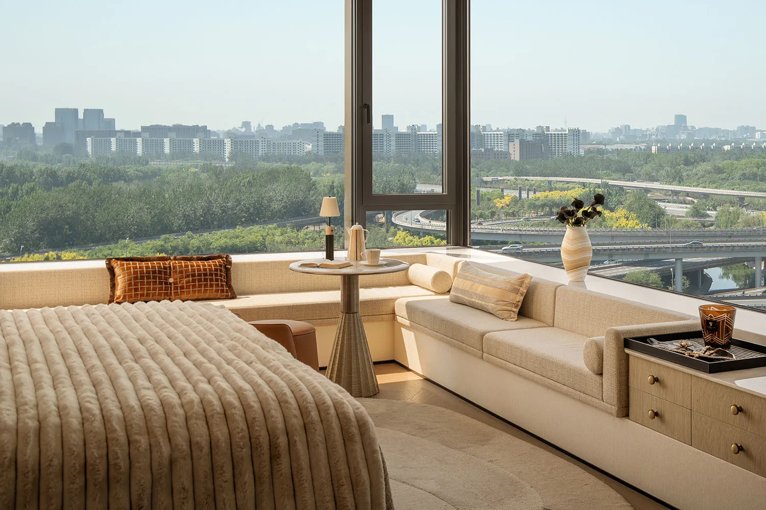 Modern high-rise bedroom featuring a large boucle fabric bed, built-in window seat with orange accent pillows, round side table, and floor-to-ceiling windows overlooking a vast cityscape with green areas and highways.